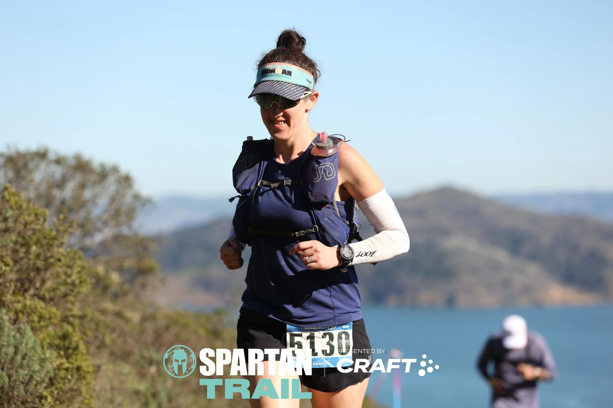 Female trail runner wearing a blue tank top, black shorts, visor, sunglasses, and a hydration pack, running outdoors near a body of water with hills in the background. The image features the Spartan Craft Trail logo.