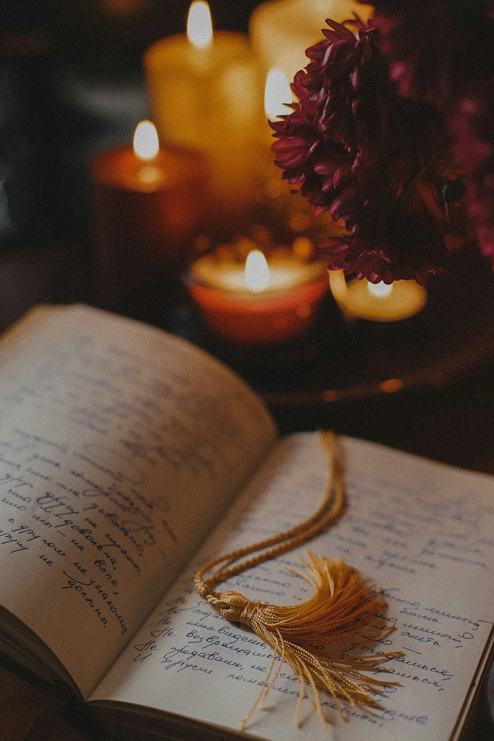 An open book written in a foreign script with a tassel bookmark, surrounded by several lit candles and a bunch of dark red flowers.