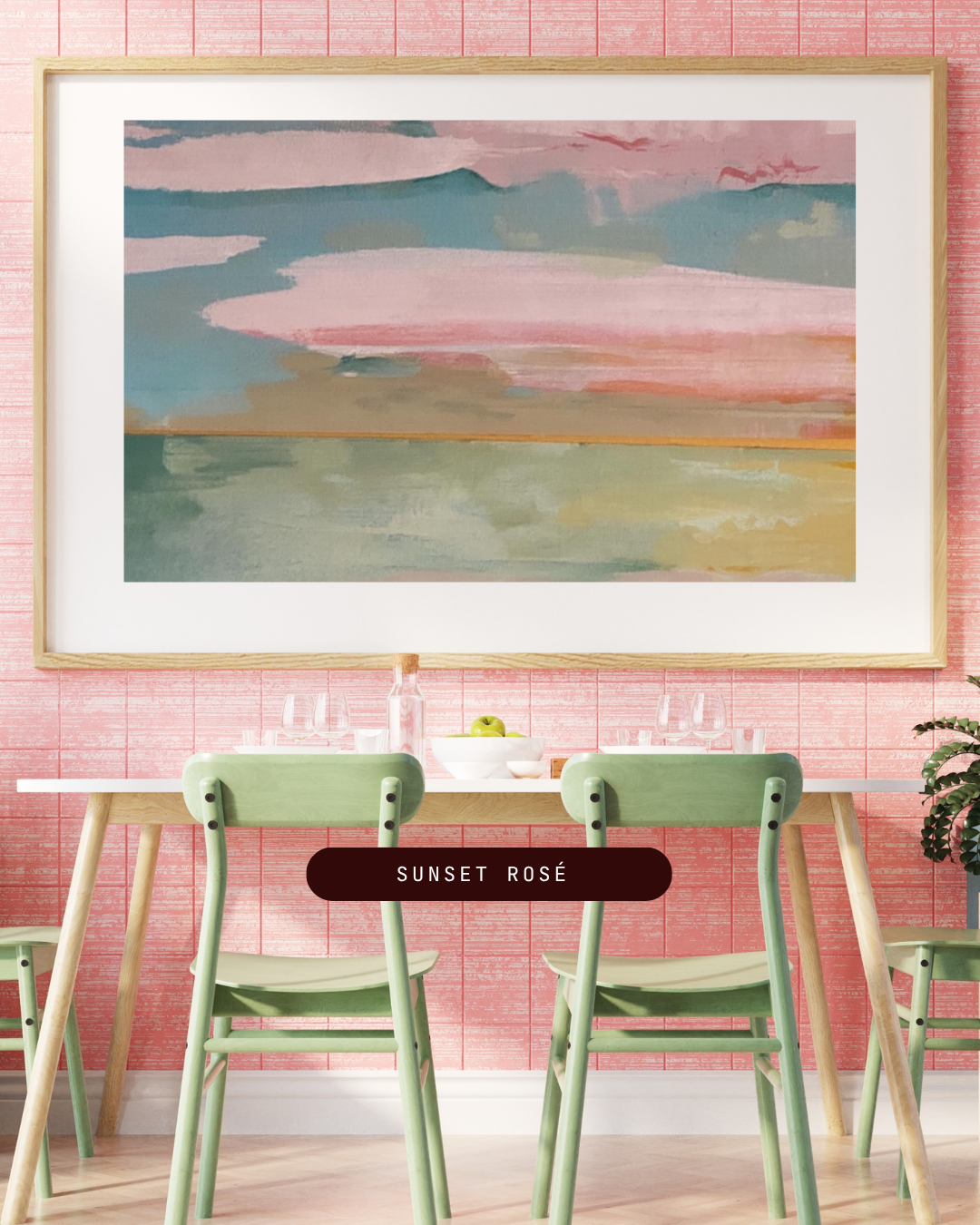 A watercolor painting of a sunset over a body of water, displayed on a pink brick wall in a dining area with green chairs and a table.