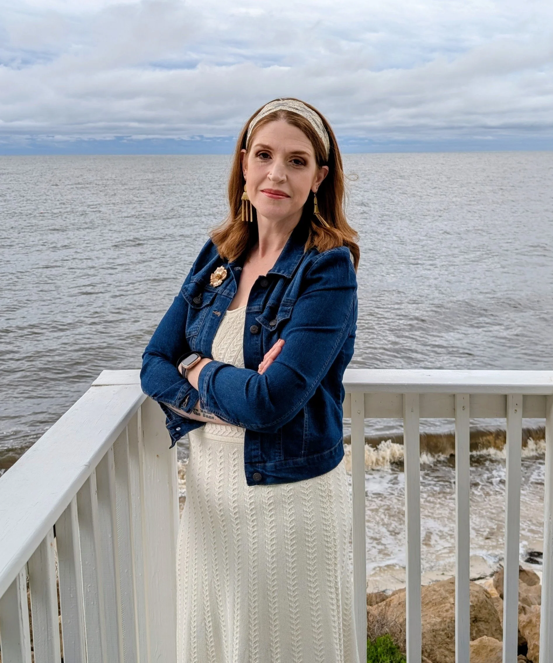 A woman with shoulder-length brown hair and earrings, wearing a denim jacket over a cream-colored sweater, standing in front of a body of water with her arms crossed. The background features an overcast sky and a railing by the water.