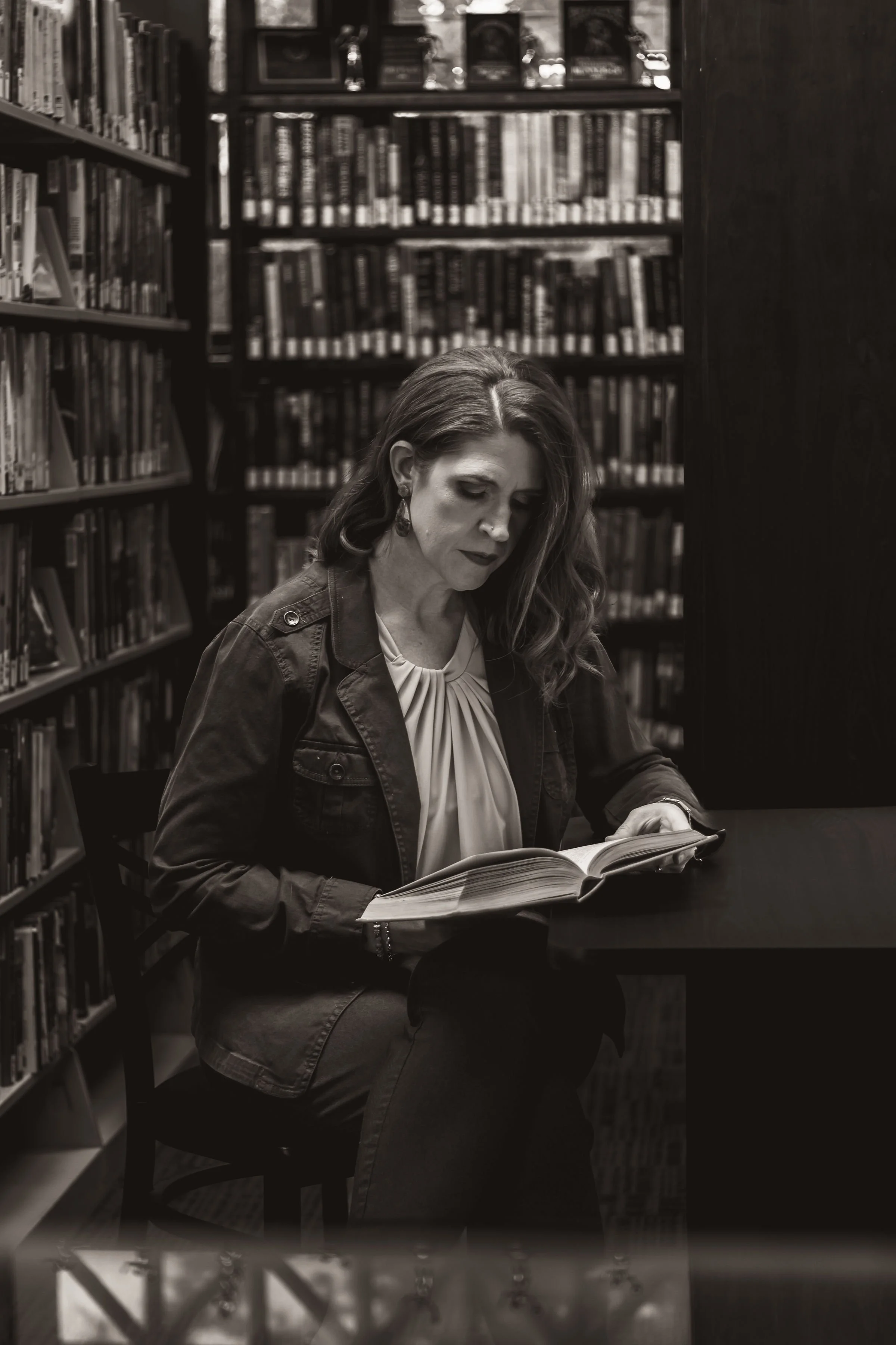 A woman with long hair and earrings sitting at a table in a library, reading a book, with shelves of books behind her.