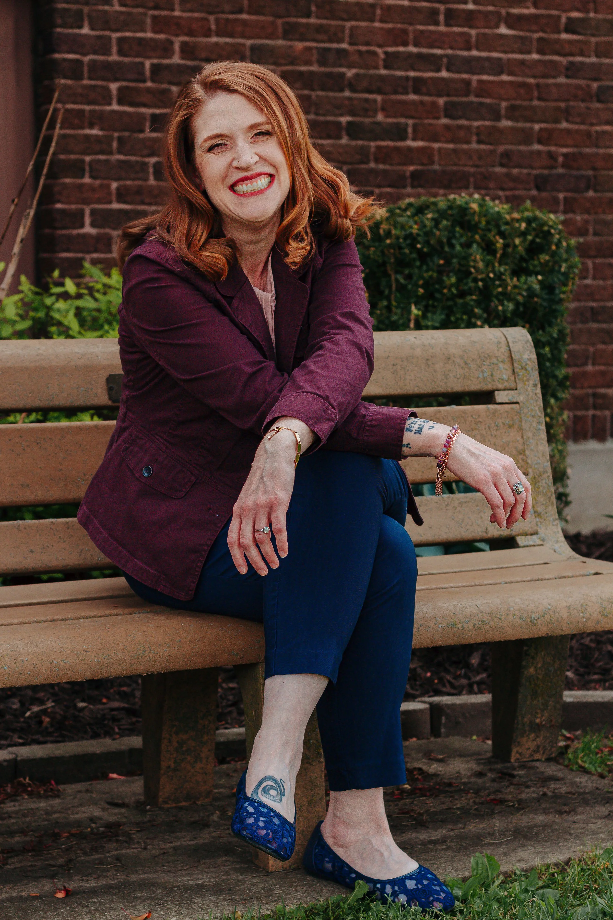 A woman with red hair wearing a maroon jacket and dark blue jeans is sitting on a park bench, smiling broadly at the camera. She has jewelry on her wrists and fingers, and is wearing blue lace-patterned shoes. The background features a brick wall and some green bushes.