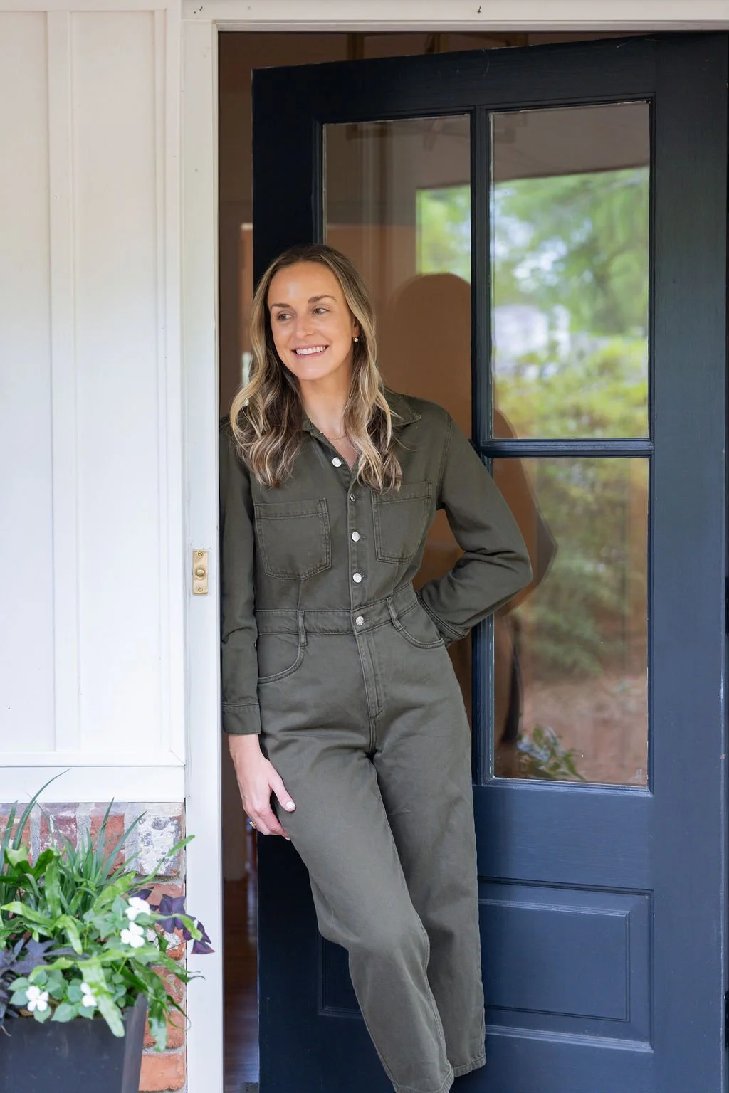 A woman with wavy blonde hair leaning against a doorframe, smiling, wearing a green jumpsuit, near a porch with a potted plant and a glass-paneled door.