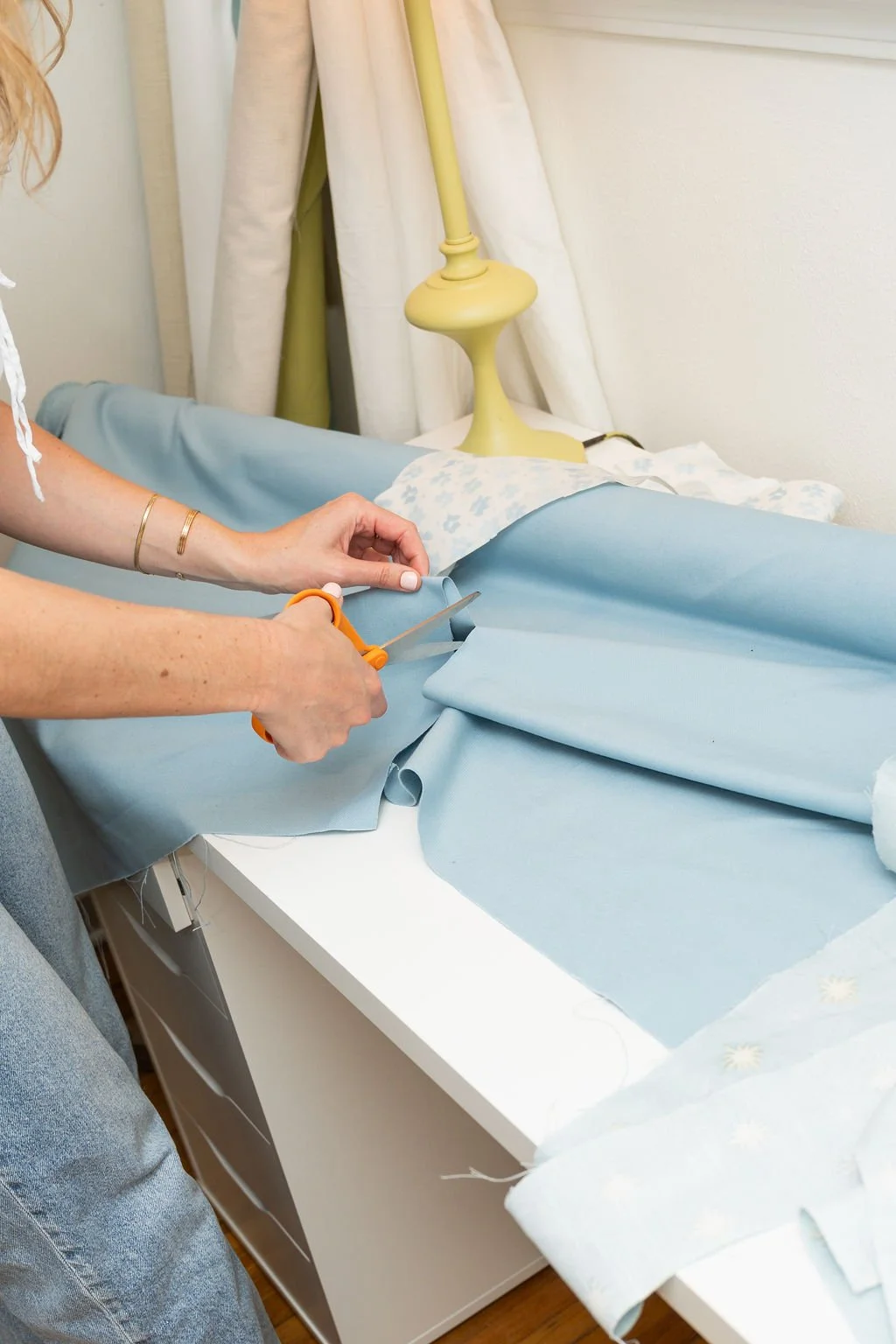 Person cutting light blue fabric with scissors on a table, with a lamp and curtains in the background.