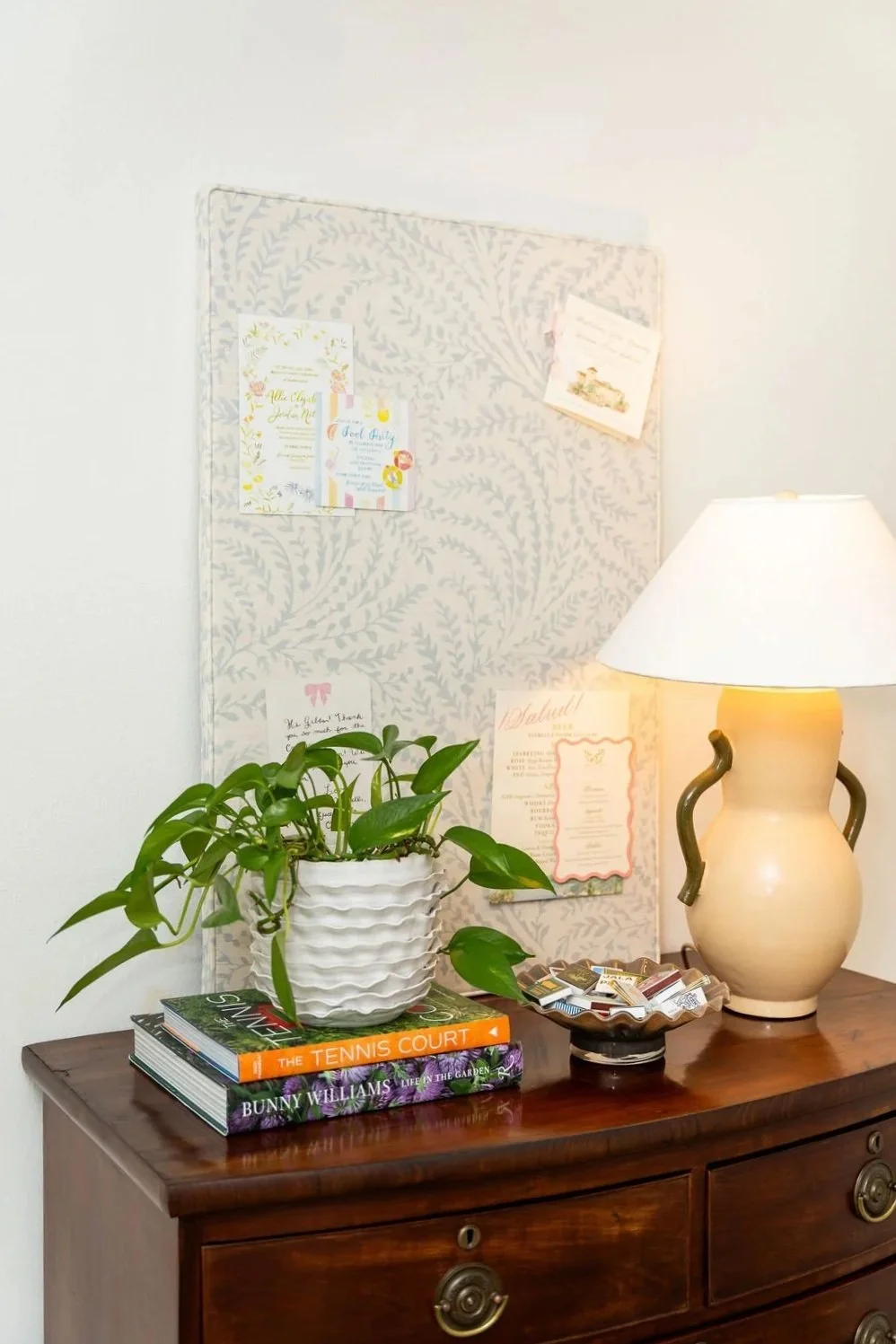 Decorative wooden sideboard with a potted plant, three stacked books, a bowl of miscellaneous small items, and a table lamp. A fabric-covered pinboard with notes and cards is mounted on the wall above.