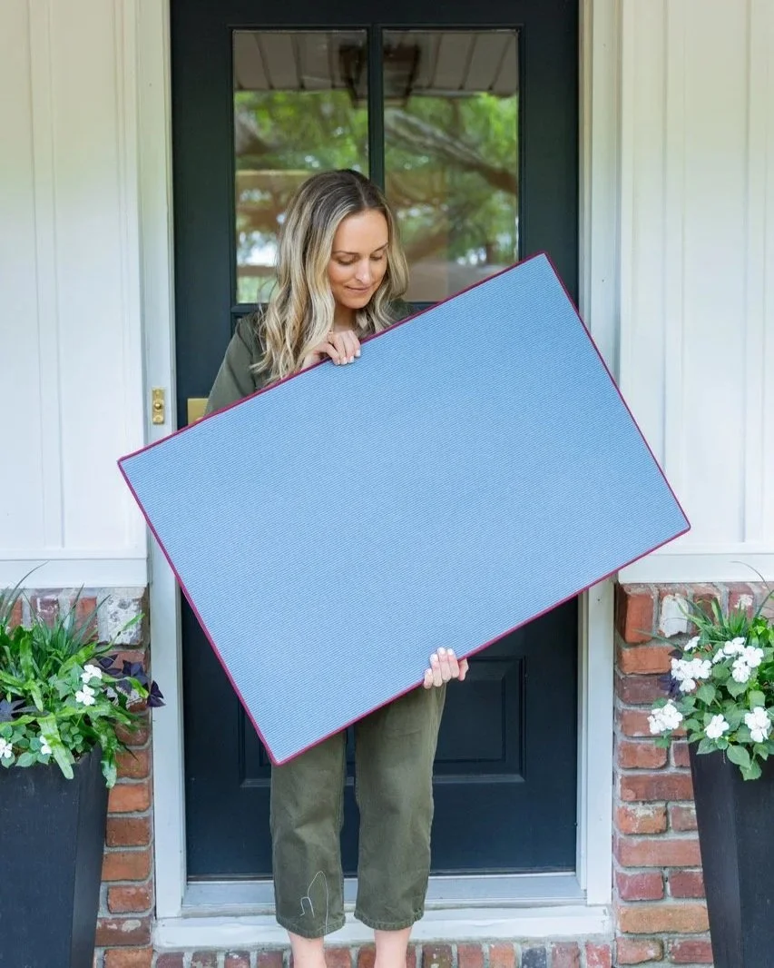A woman standing on a porch holding a large, blank, gray doormat with a red border in front of a black door with a window. There are two large planters with white flowers and green leaves on either side of her.
