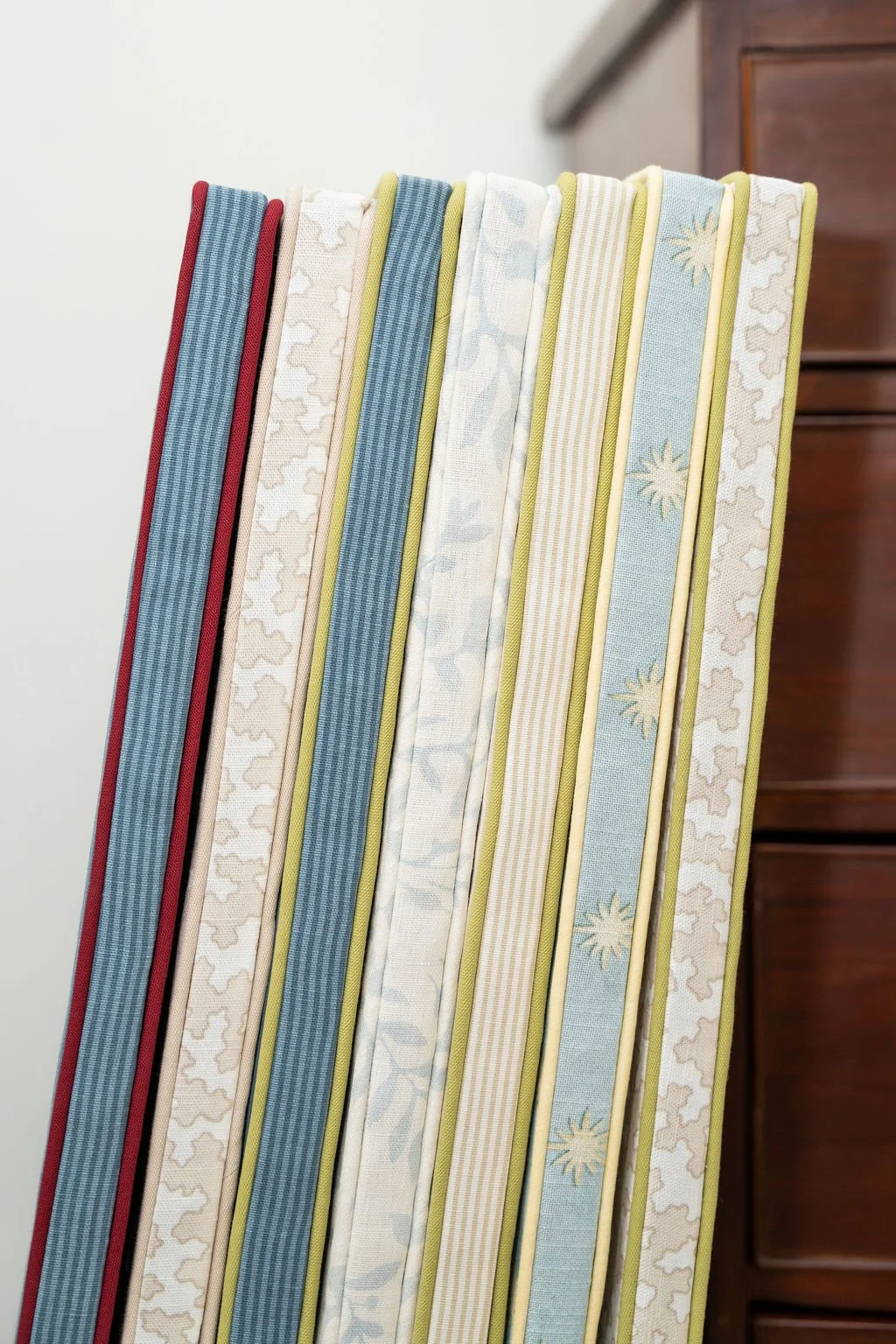 A vertical stack of eight different fabric-covered mattresses or cushions with various patterns and colors, placed next to a brown wooden piece of furniture.