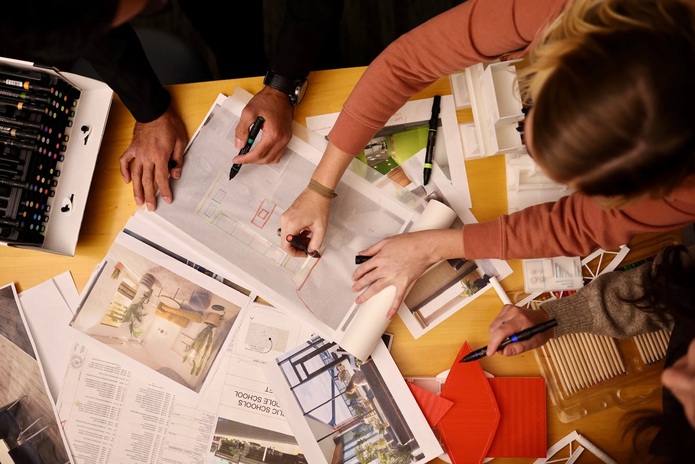 Two individuals working on a design project at a table covered with plans, sketches, and printed images, with writing utensils and color markers.