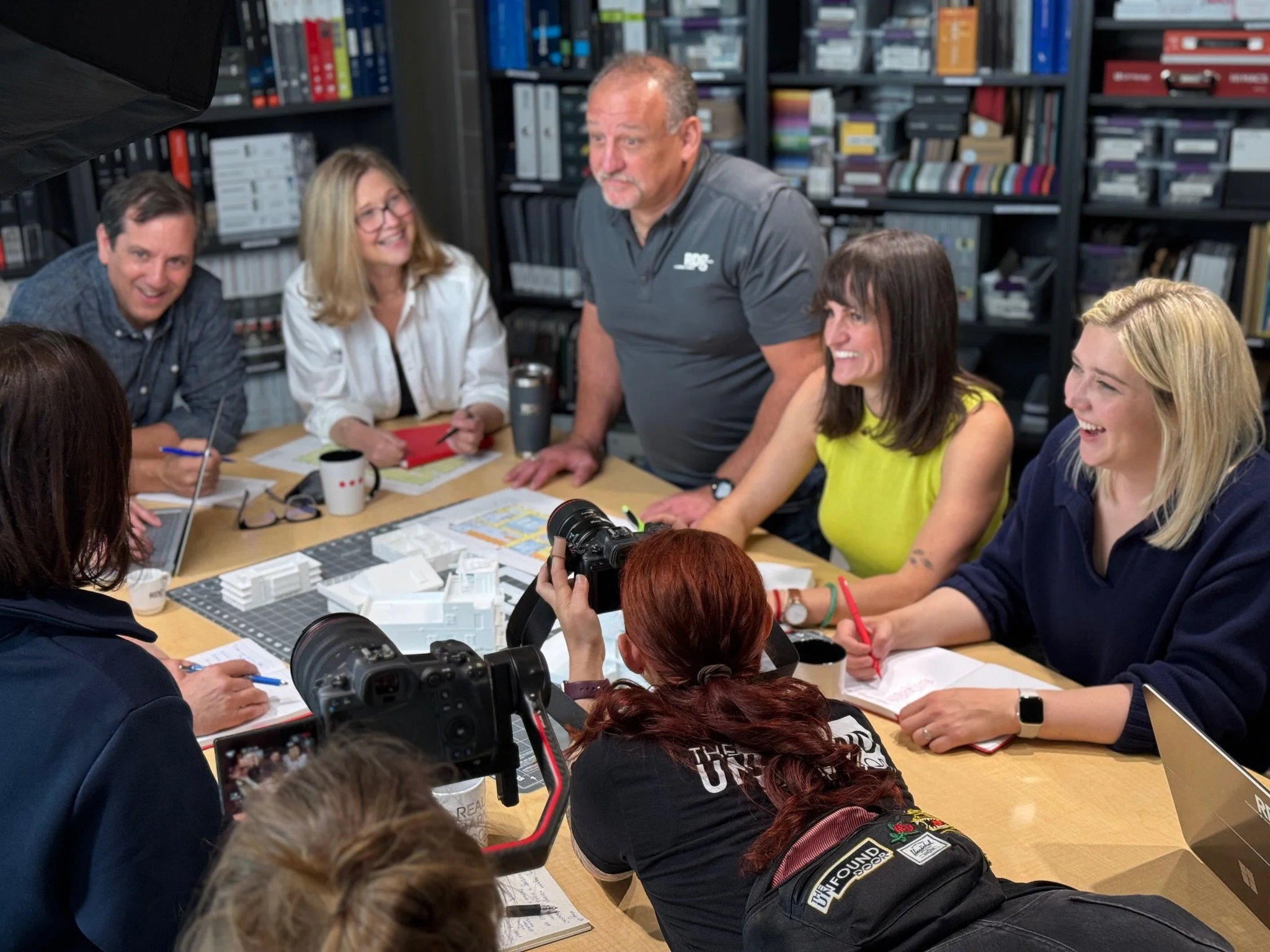 A diverse group of six adults gathered around a table in a meeting room, engaged in a discussion. They are smiling and appear to be collaborating, with some taking notes, and a camera is capturing the moment. The room has shelves filled with books an