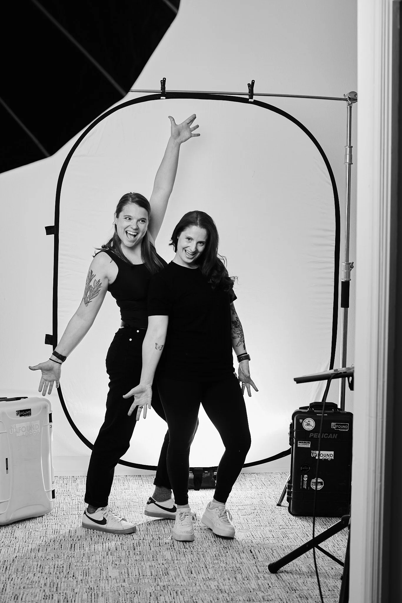 Two women posing playfully in a photography studio with a white backdrop, smiling and showing relaxed body language.