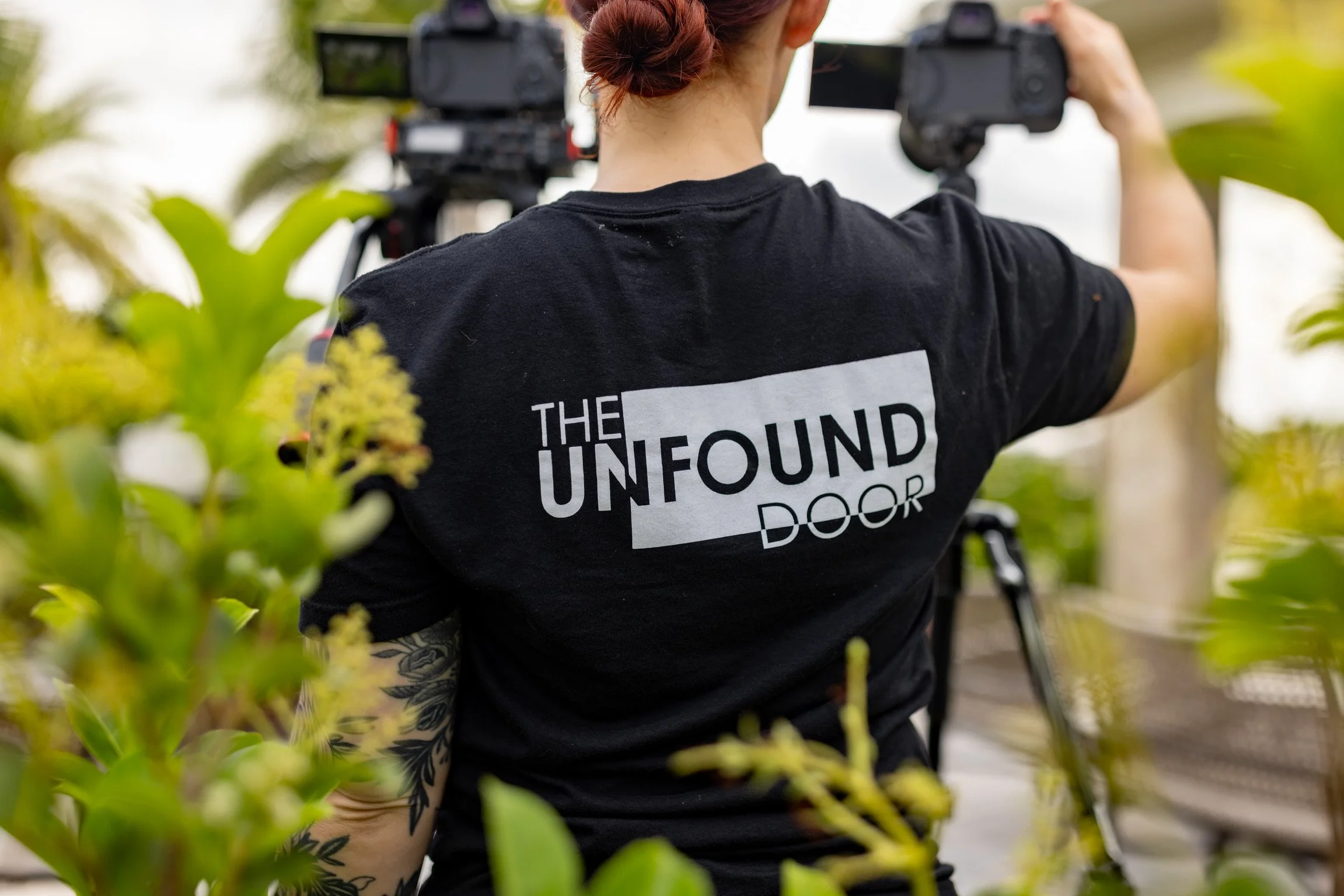 Person with red hair in a bun wearing a black T-shirt with the logo 'THE UNFOUND DOOR' on the back, operating a video camera outdoors among green plants.