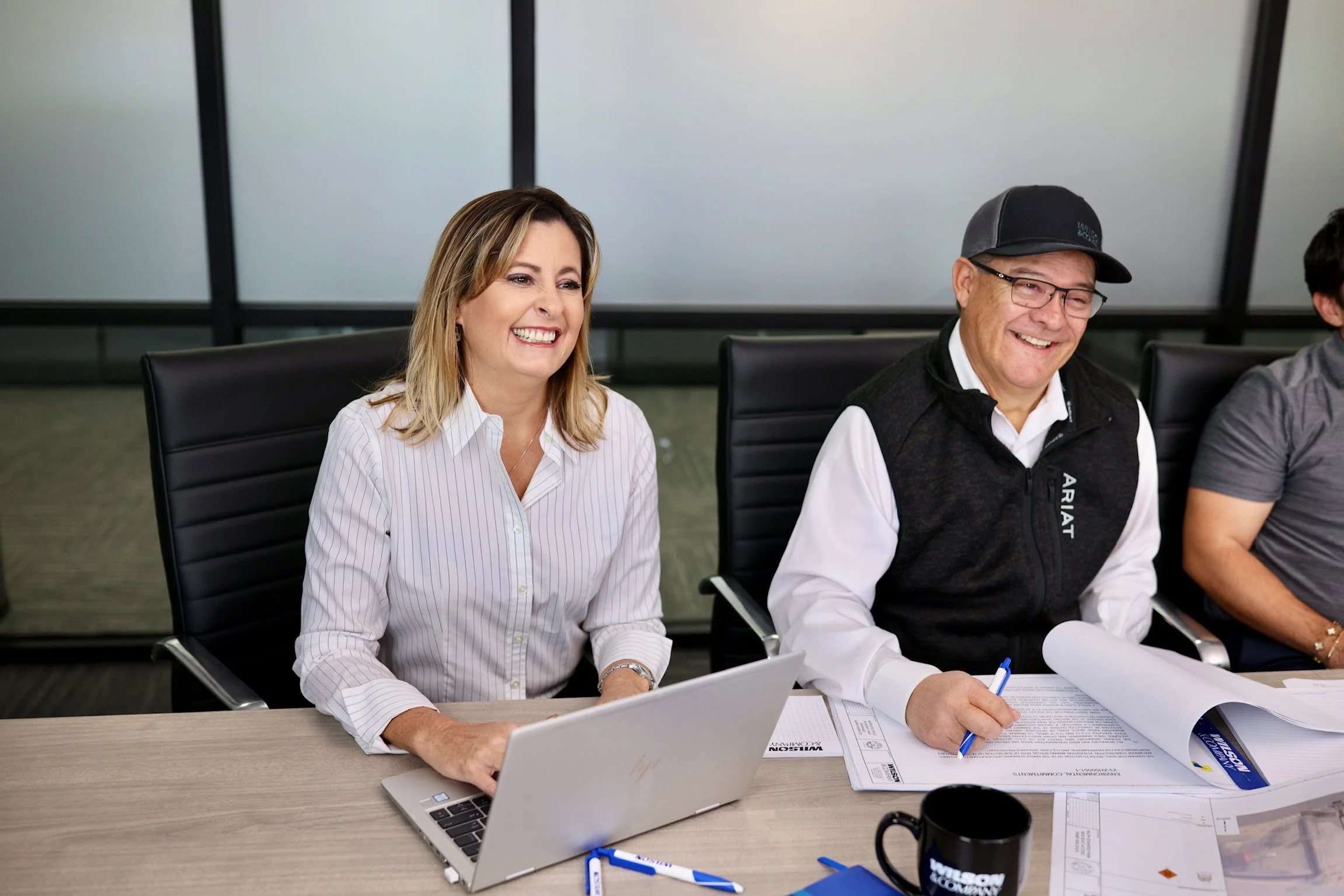 Smiling woman with shoulder-length blonde hair in a white striped shirt sitting at a table with a silver laptop, and a man with glasses, a black vest, and a black cap sitting next to her writing on documents in a conference room.