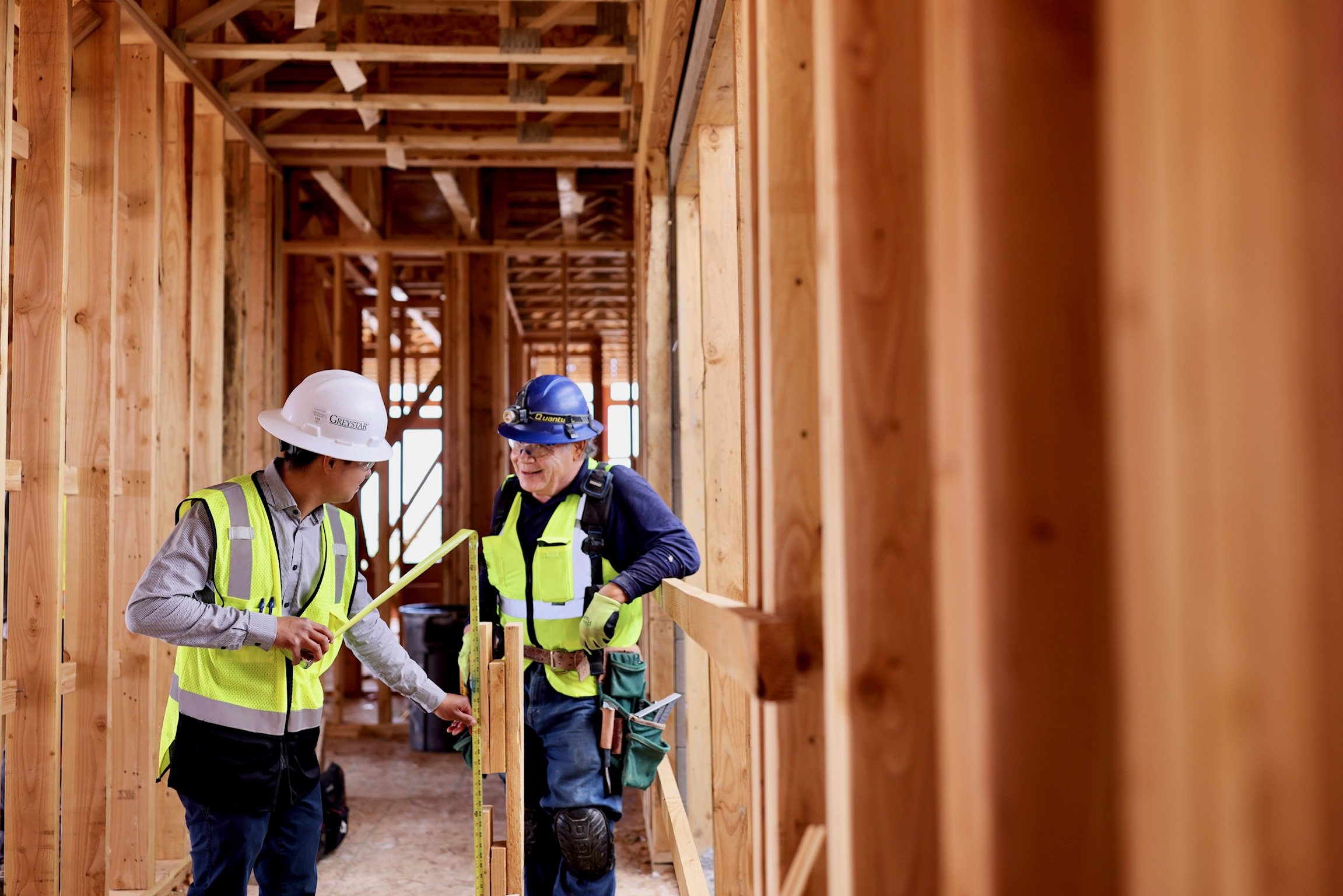 Two construction workers, wearing safety helmets and reflective vests, are working inside a wooden framing structure of a building under construction. They are examining or measuring something with a yellow measuring tape.
