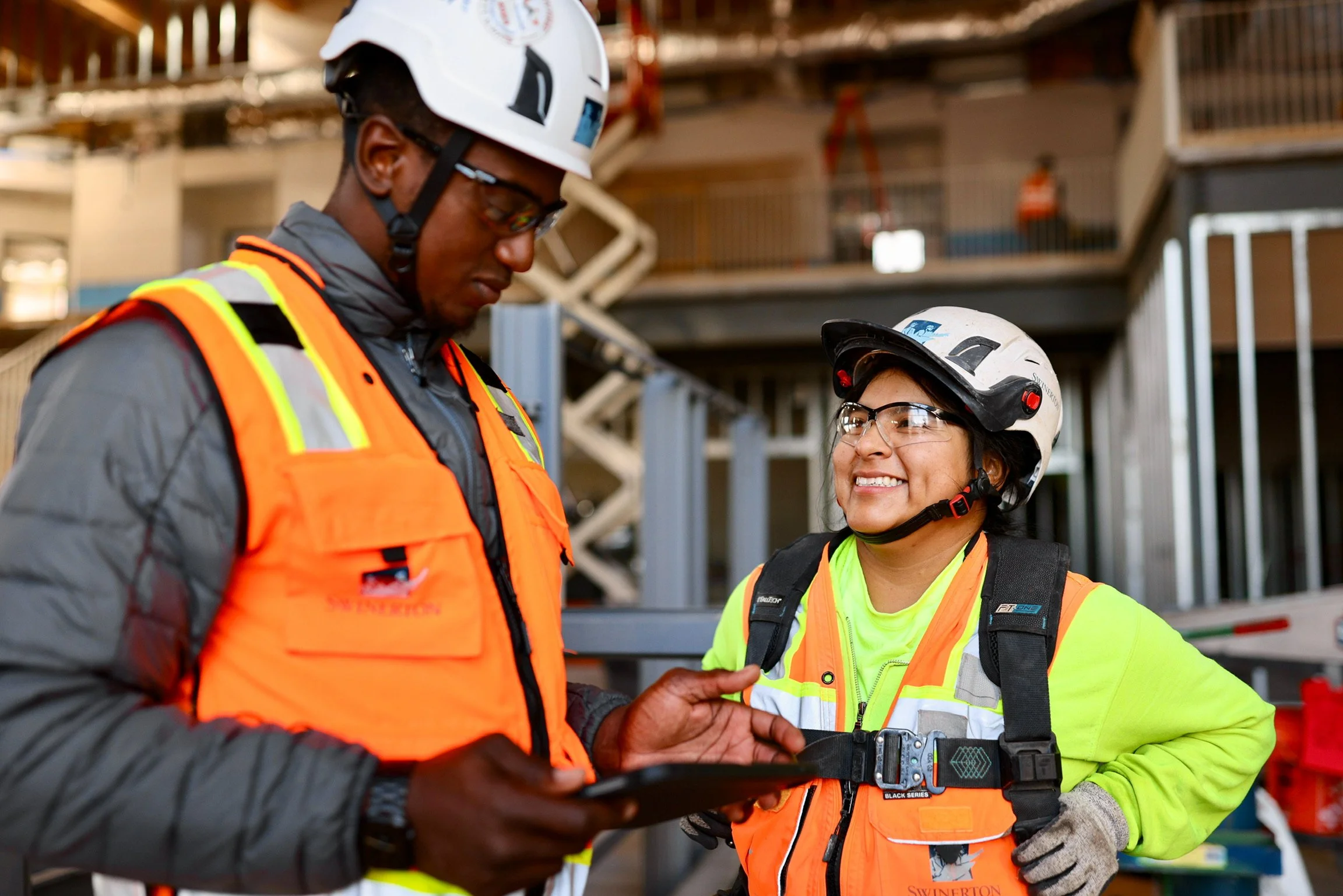 Two construction workers wearing safety helmets, high-visibility vests, and safety glasses are engaged in a conversation inside a construction site with construction equipment visible in the background.