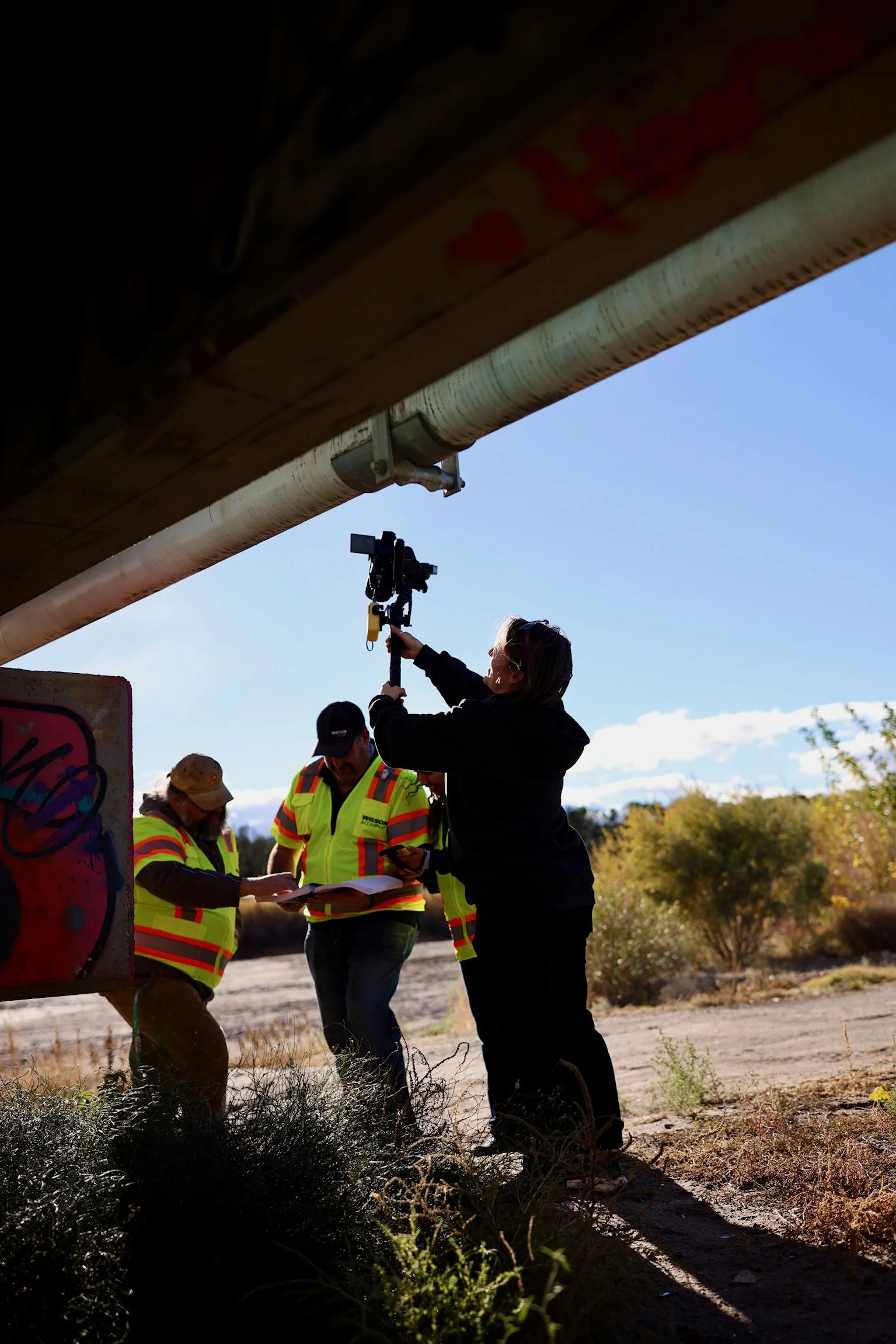 People working outdoors under a bridge, with one person recording on a camera and two others examining papers, wearing high-visibility safety vests.