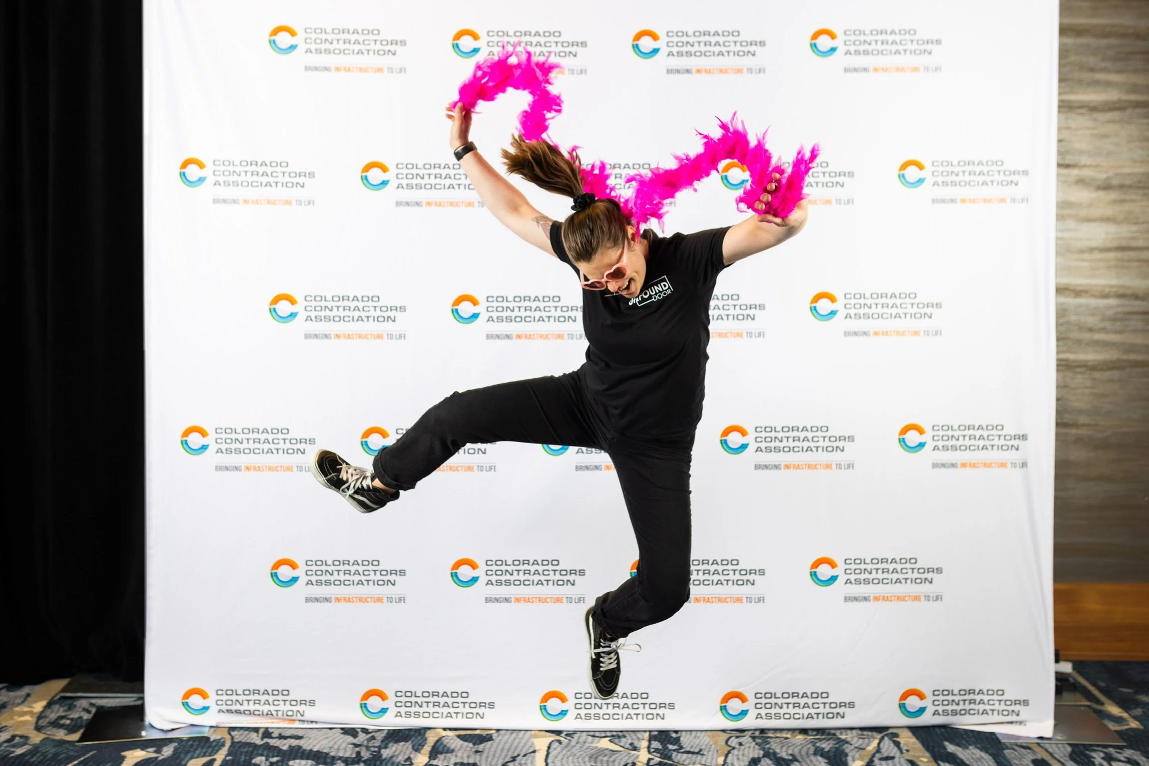 Person jumping with pink feather boas in front of a Colorado Contractors Association backdrop.