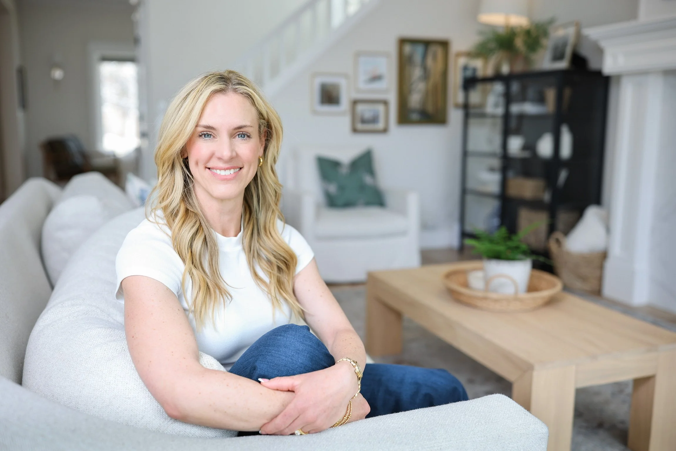 A smiling woman with blonde hair, sitting on a light-colored couch in a cozy, well-decorated living room with framed pictures on the wall and houseplants.