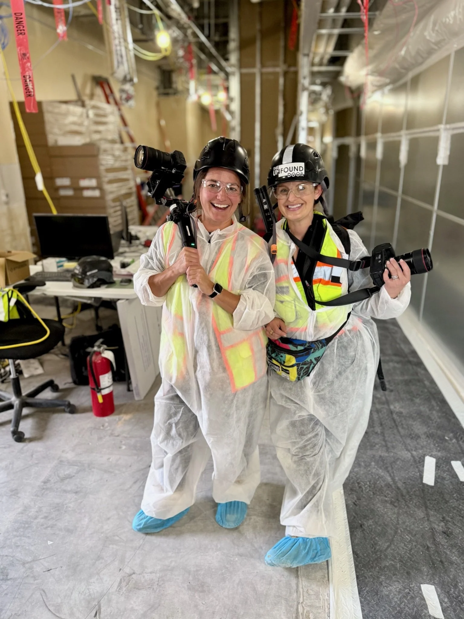 Two women in protective gear holding cameras on a construction site, smiling for the photo.