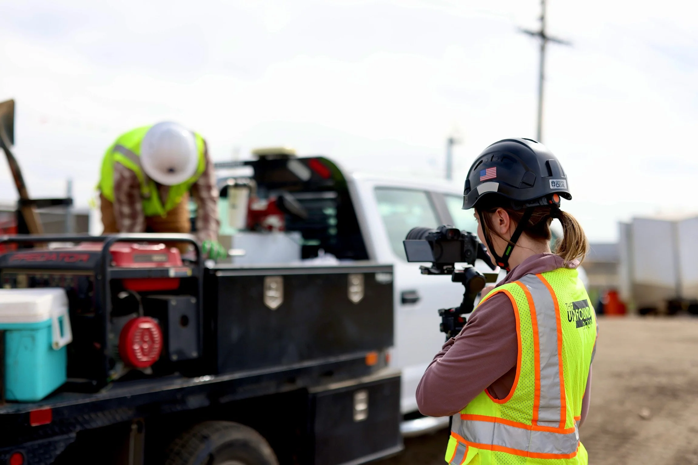 A woman wearing a safety helmet and a fluorescent safety vest operating a camera on a tripod outdoors, with a utility worker in a hard hat and high visibility vest working near a utility truck.