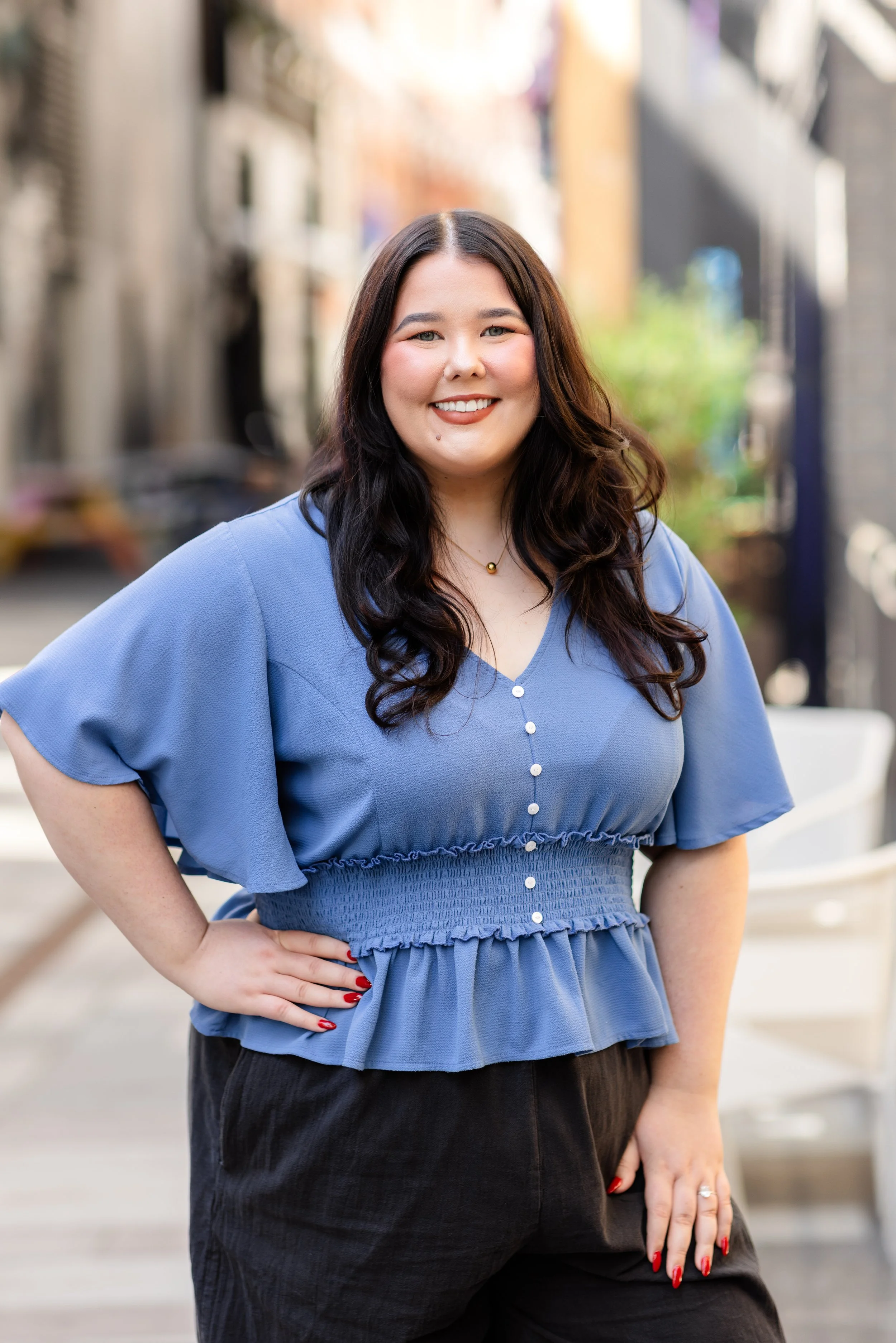 A woman with long, dark hair and fair skin smiling outdoors, wearing a blue blouse with buttons and black pants.