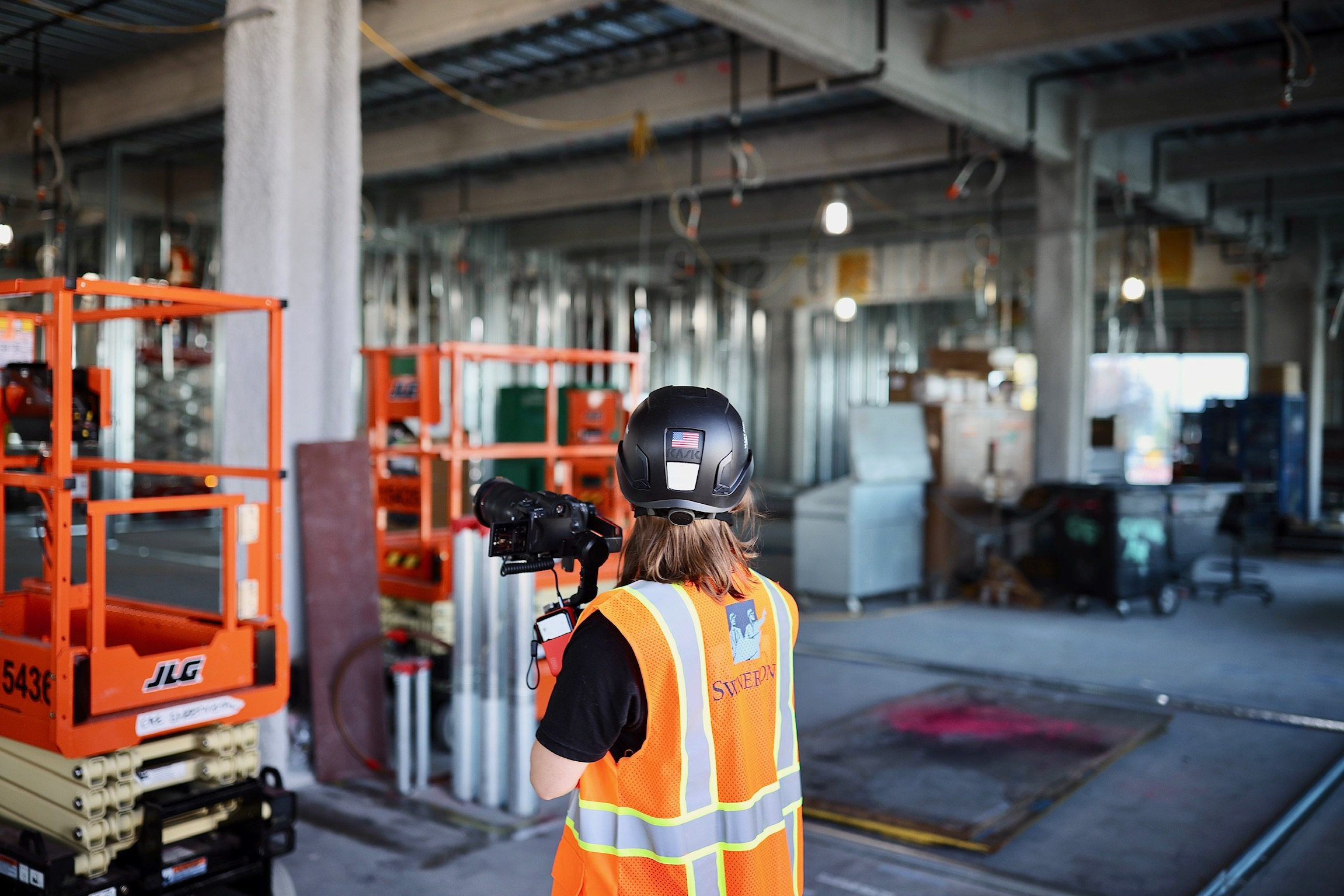 A construction worker in a black helmet and orange safety vest holding a camera inside an unfinished building with construction equipment and materials.