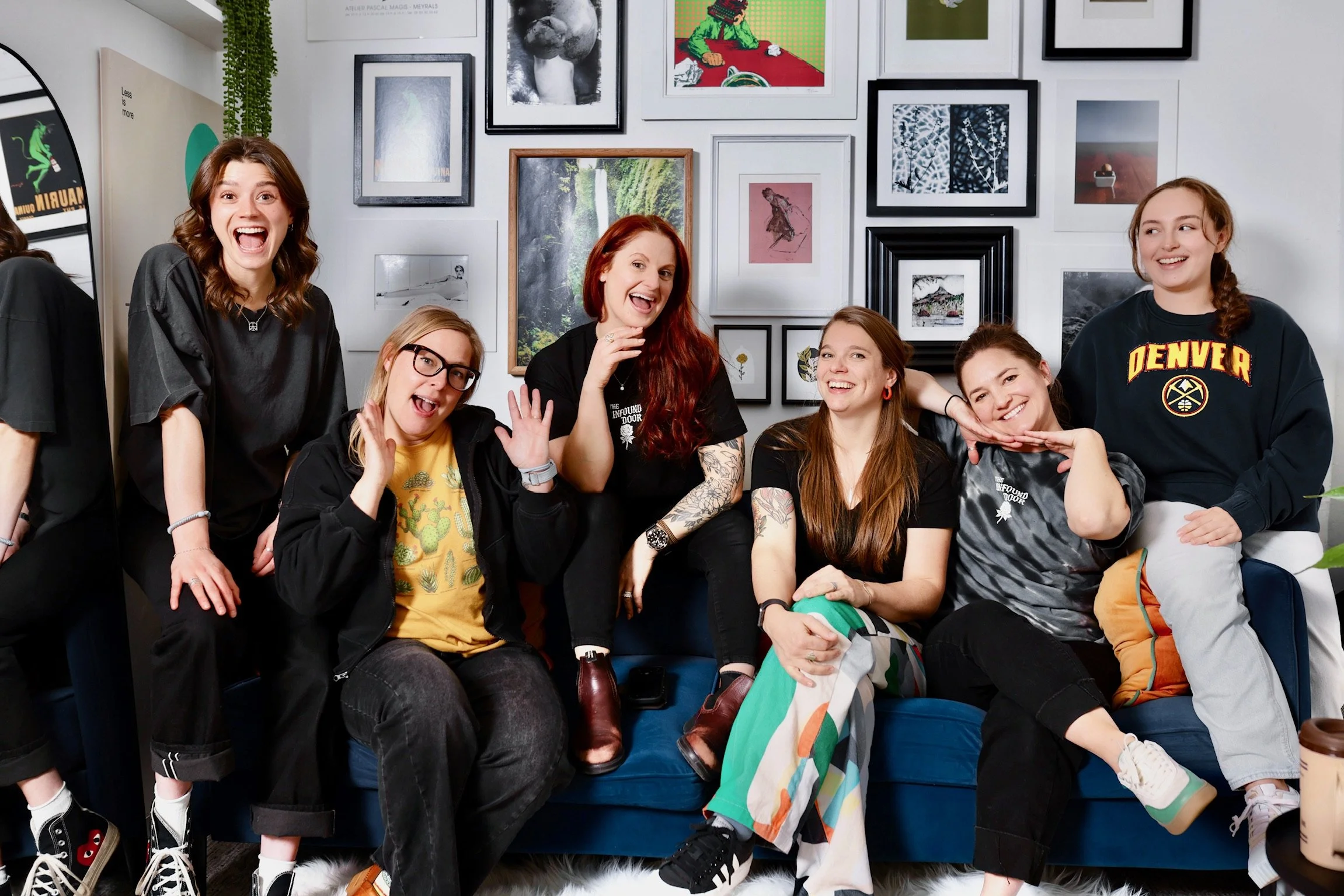 Group of seven young women sitting and standing in a living room with a gallery wall of framed artwork behind them, smiling and posing for the photo.
