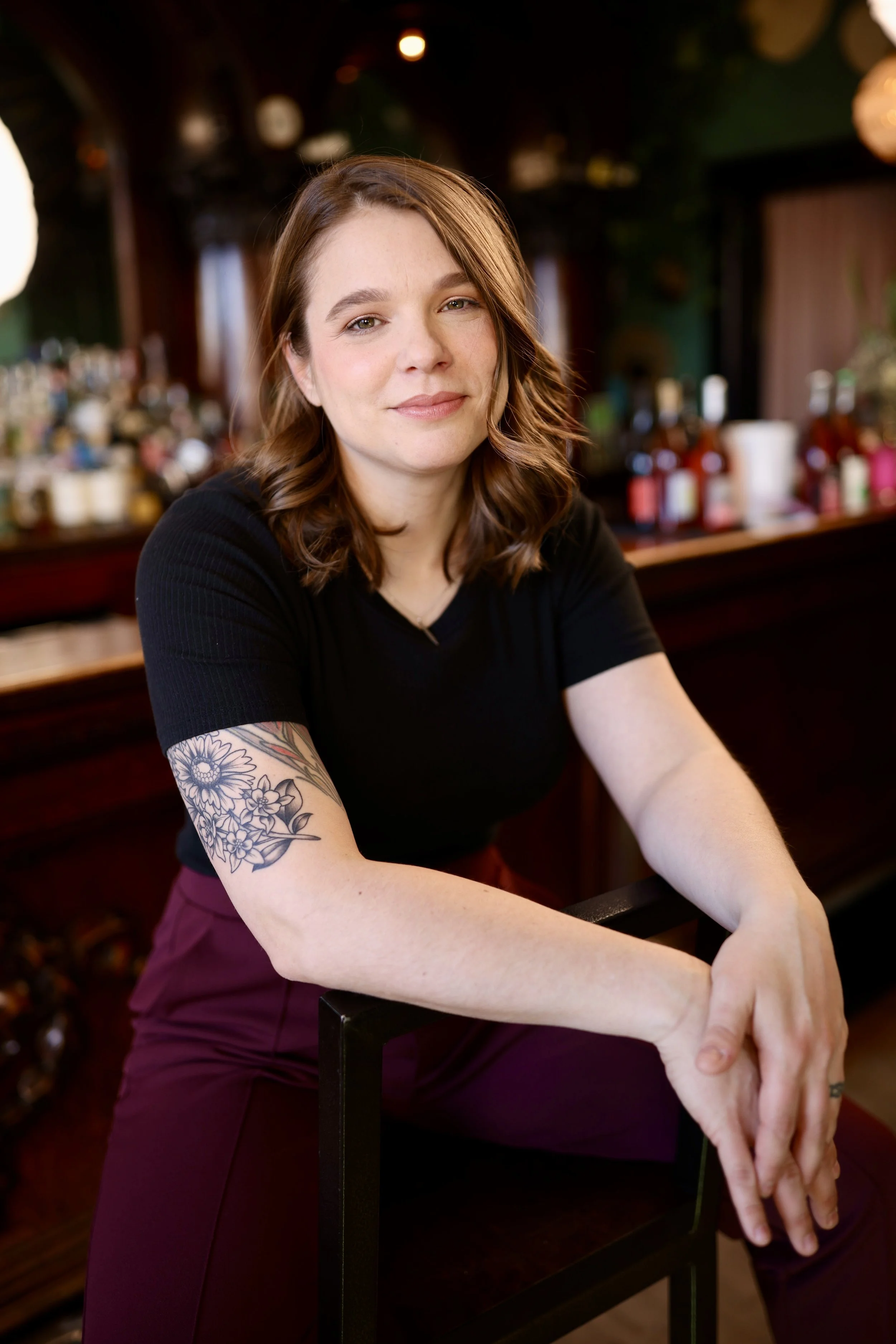 A woman with shoulder-length brown hair, wearing a black top and burgundy pants, sitting at a bar with blurred bottles and bar counter in the background.