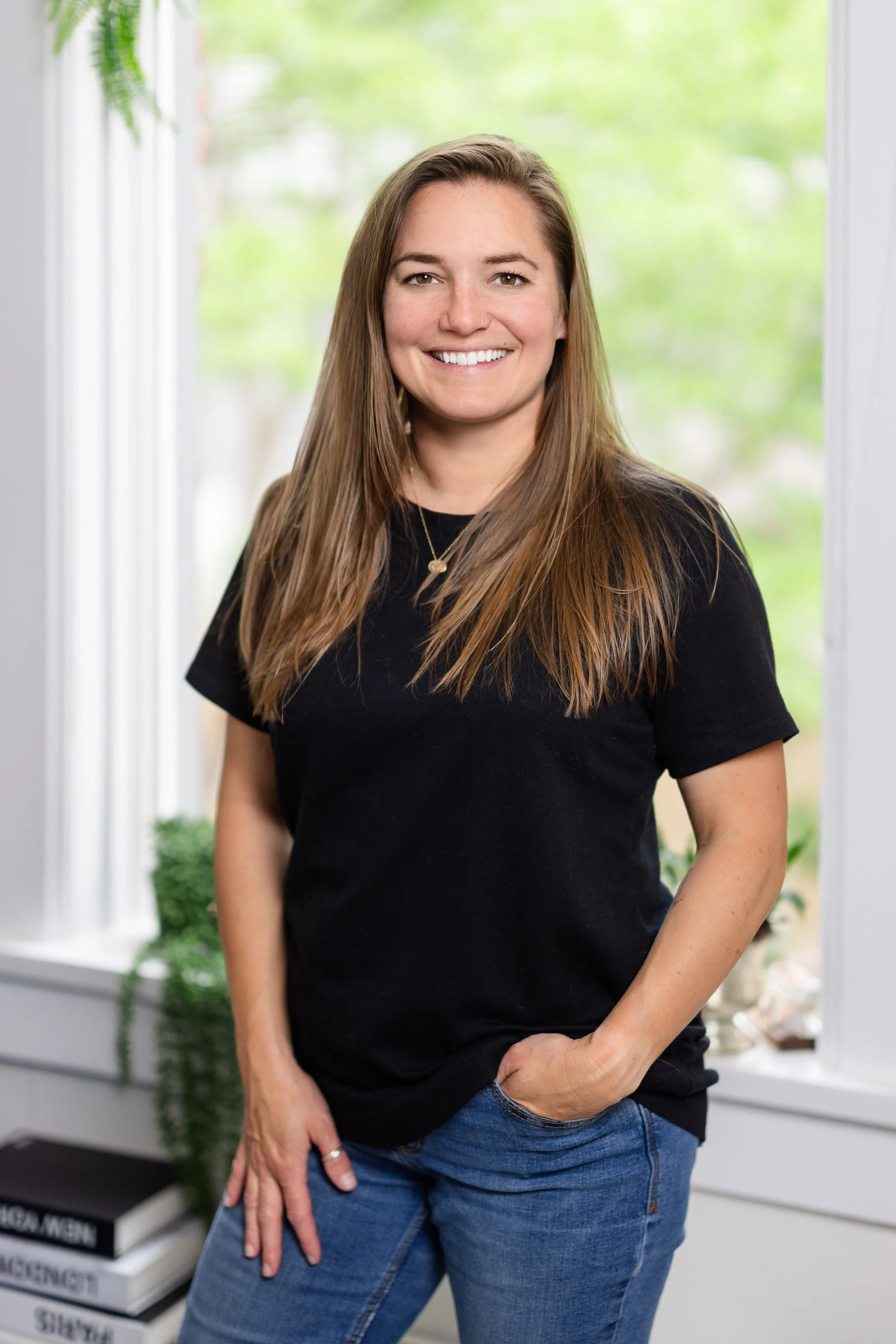 A woman with long, straight, light brown hair is smiling and wearing a black t-shirt and blue jeans. She is standing inside near a window with a green outdoor background.