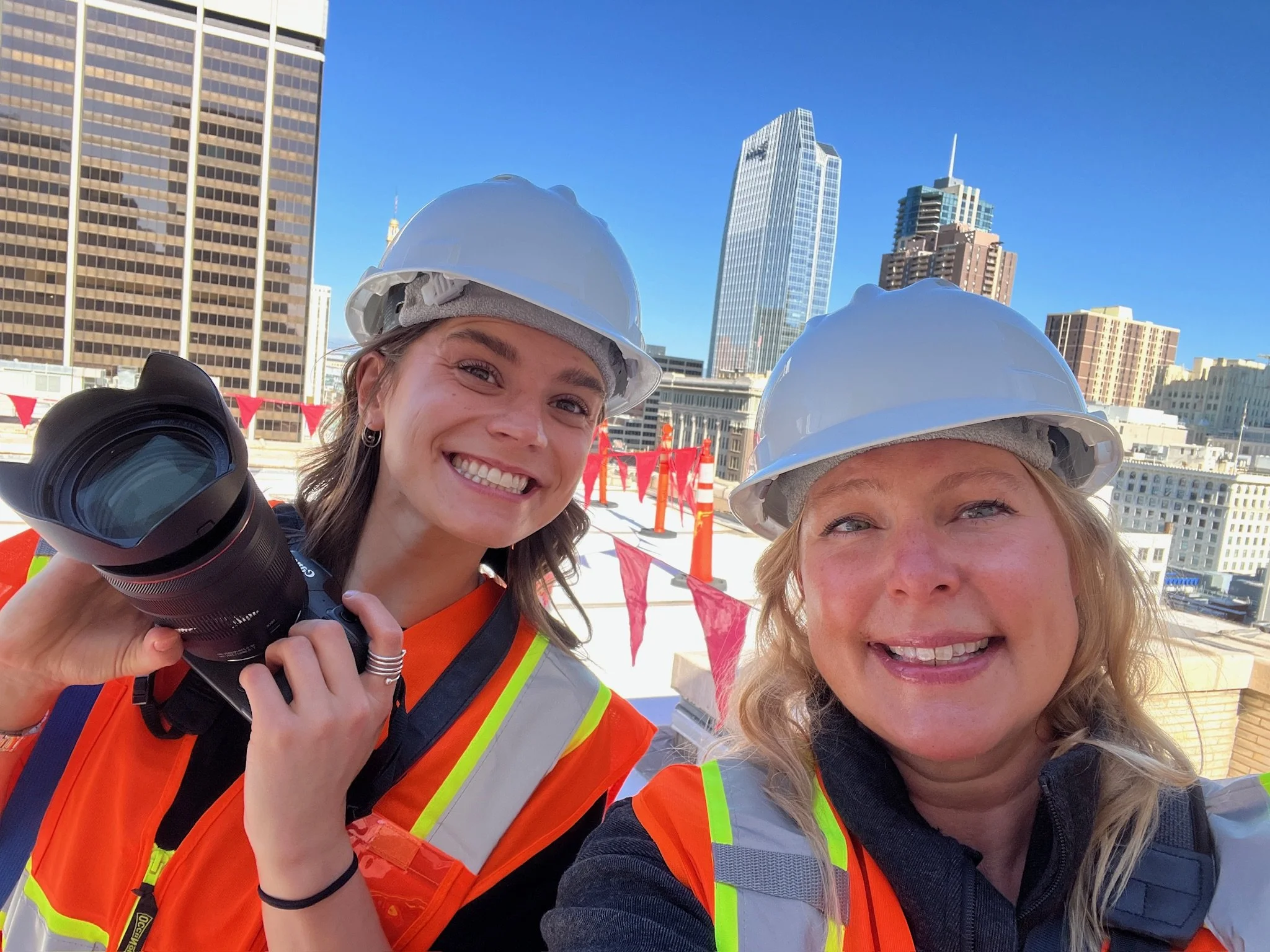 Two women wearing safety helmets and orange safety vests taking a selfie on a rooftop with city skyscrapers in the background.
