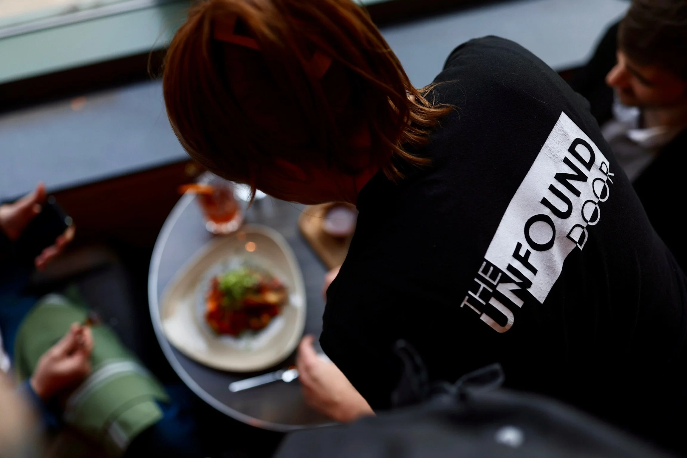 Overhead view of a person with red hair wearing a black hoodie that says 'THE UNIFORM DOOR' on the back, sitting at a table with a plate of food and a glass of drink, surrounded by other people.
