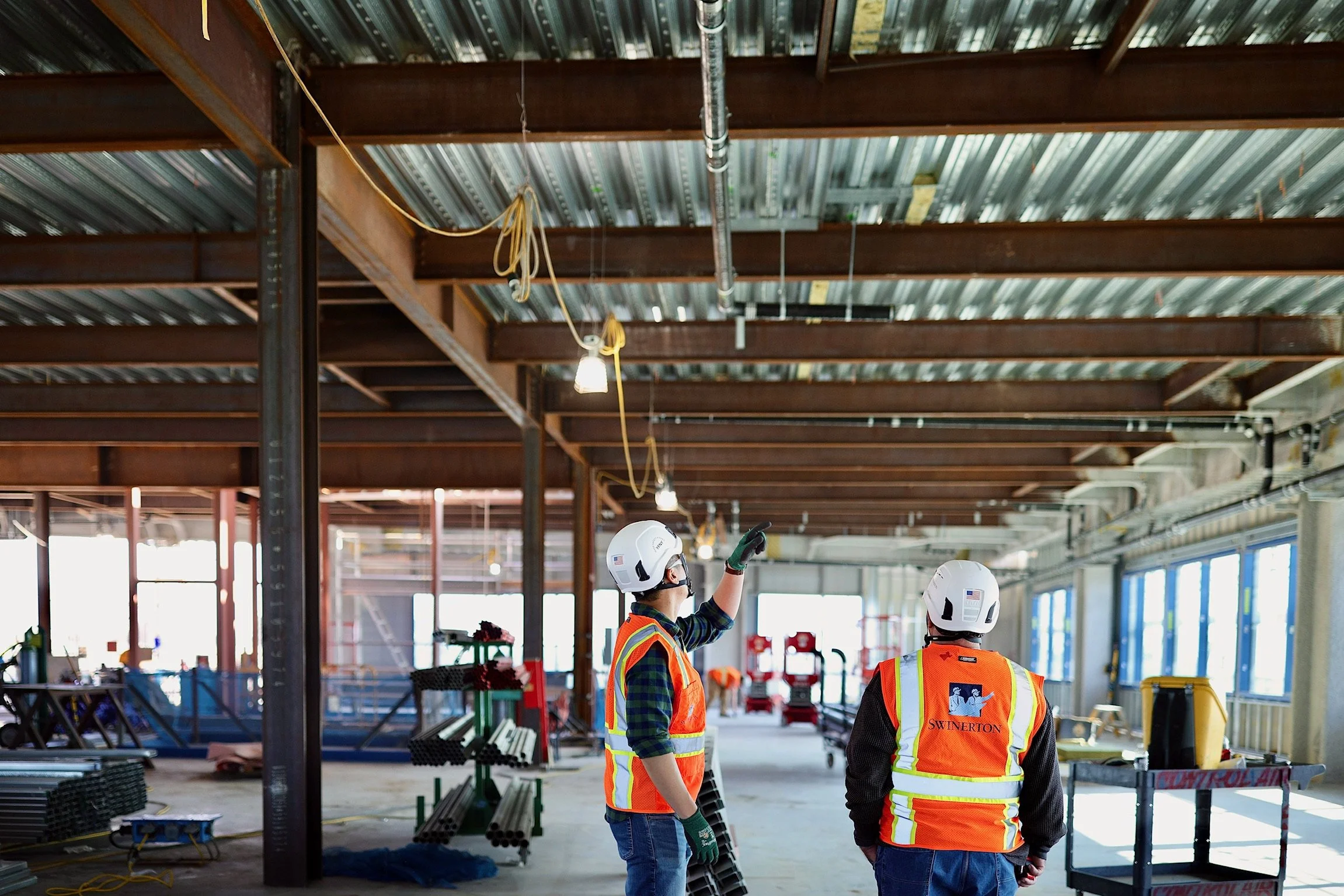 Construction workers in safety vests and helmets inside a building under construction, with exposed metal beams and construction materials around.