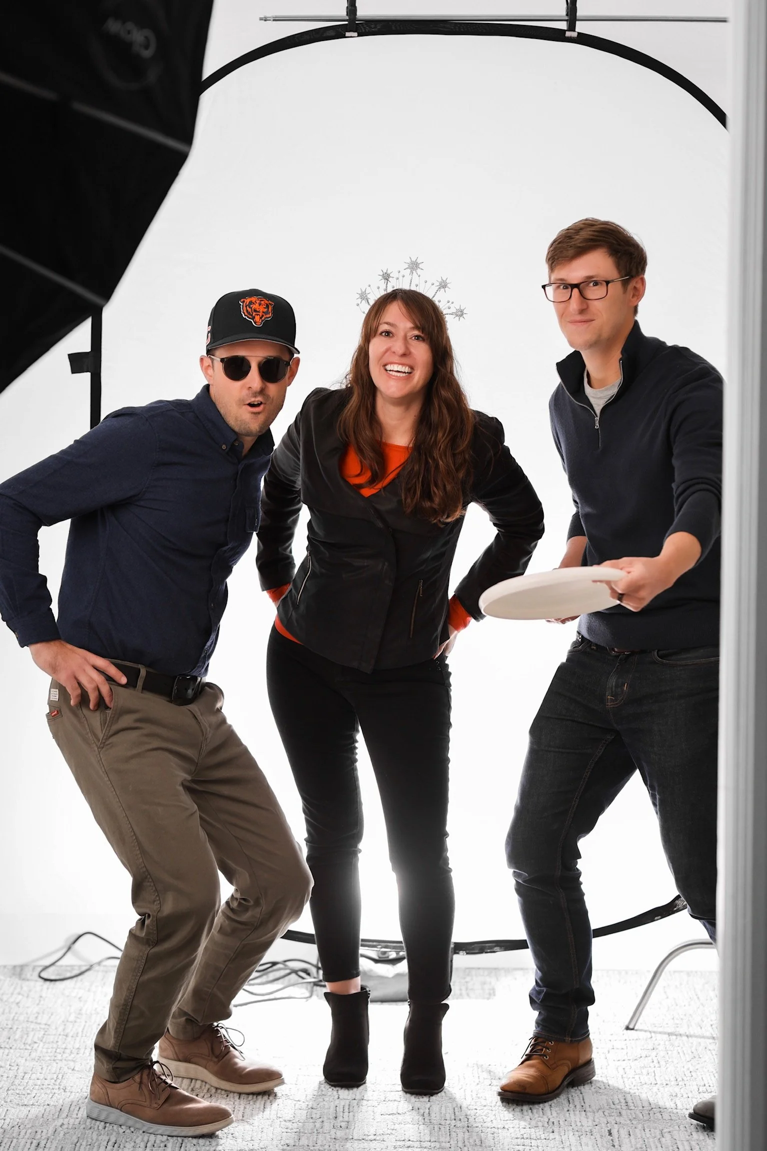 Three people standing inside a photography studio with a plain white background, posing for a photo. The person in the middle has a snowflake halo headband, the person on the left is wearing sunglasses and a Chicago Bears cap, and the person on the right is holding a white plate.
