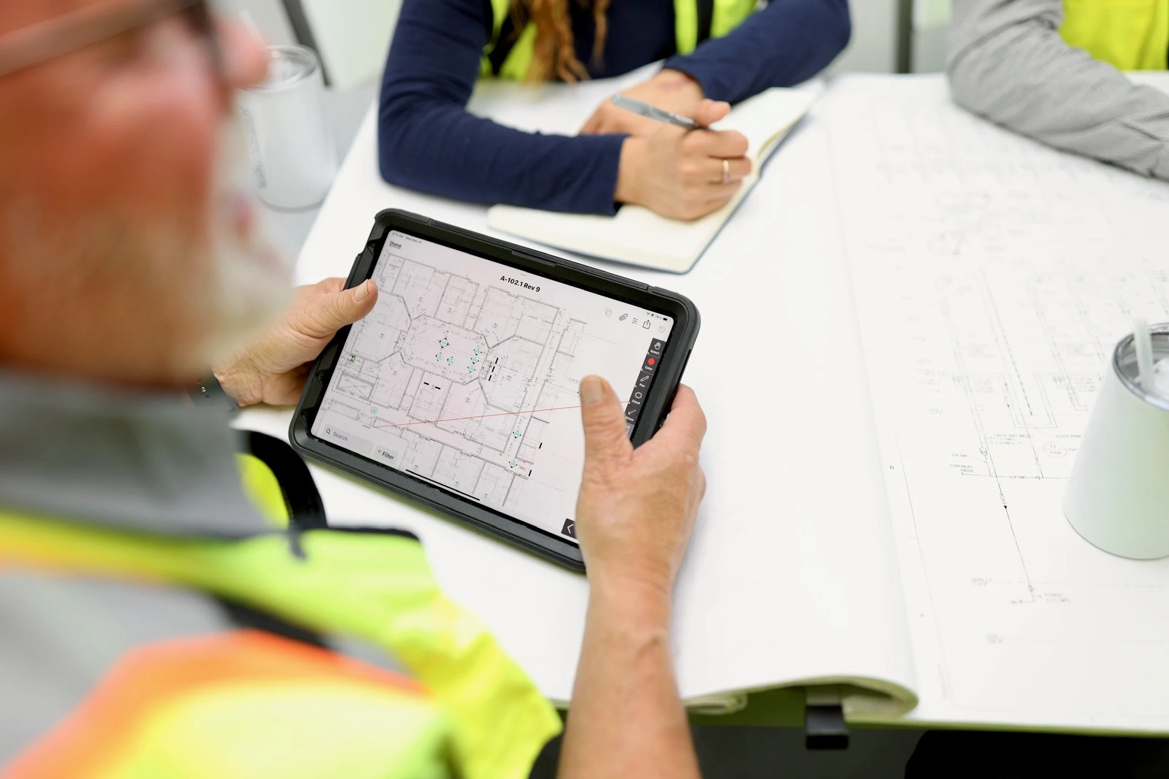 Person holding a tablet displaying a floor plan while others review architectural blueprints on a table during a meeting.