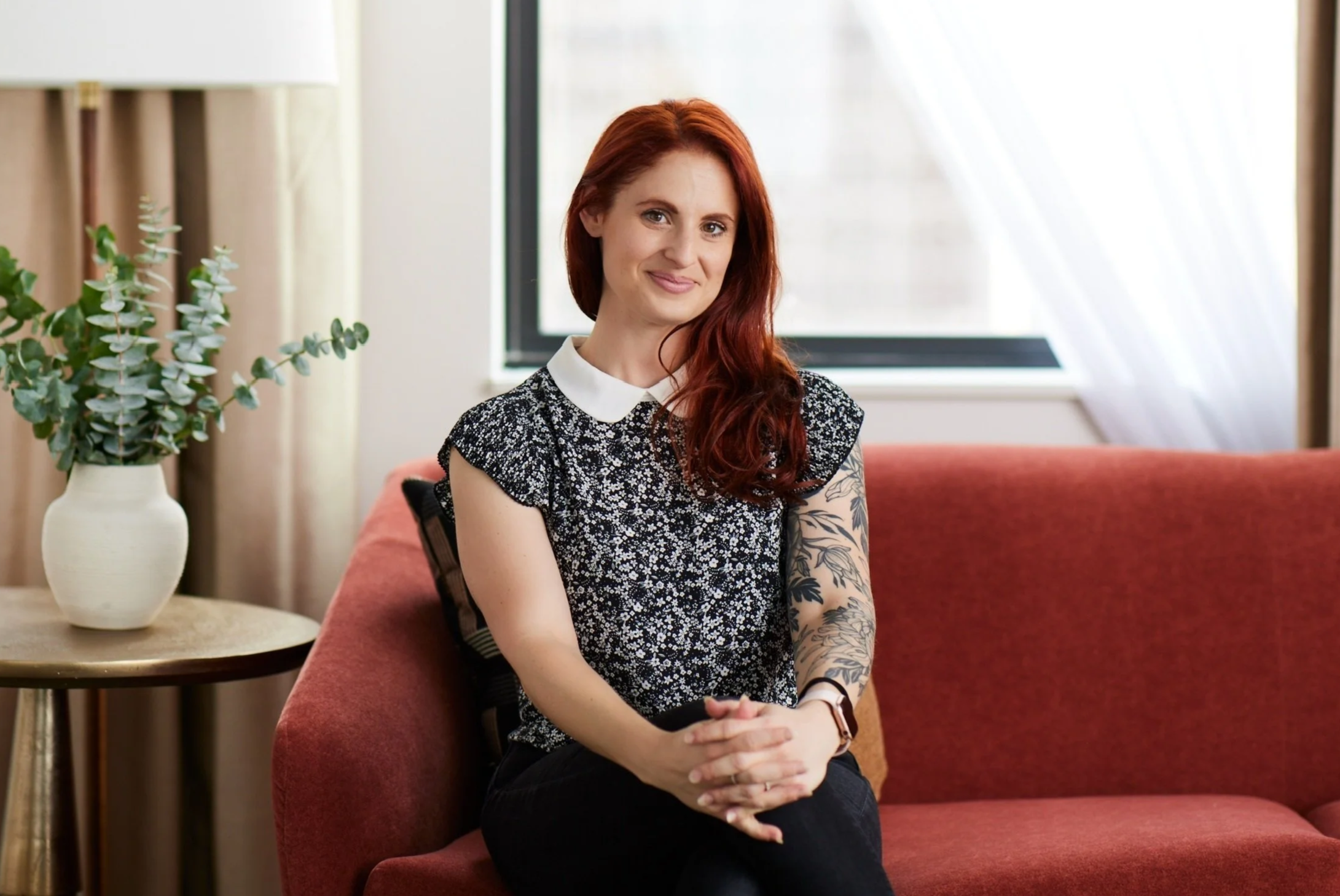 A woman with red hair and tattoos on her arm sitting on a red sofa in a bright room with a window, a white vase with greenery on a small table, and cream-colored curtains.