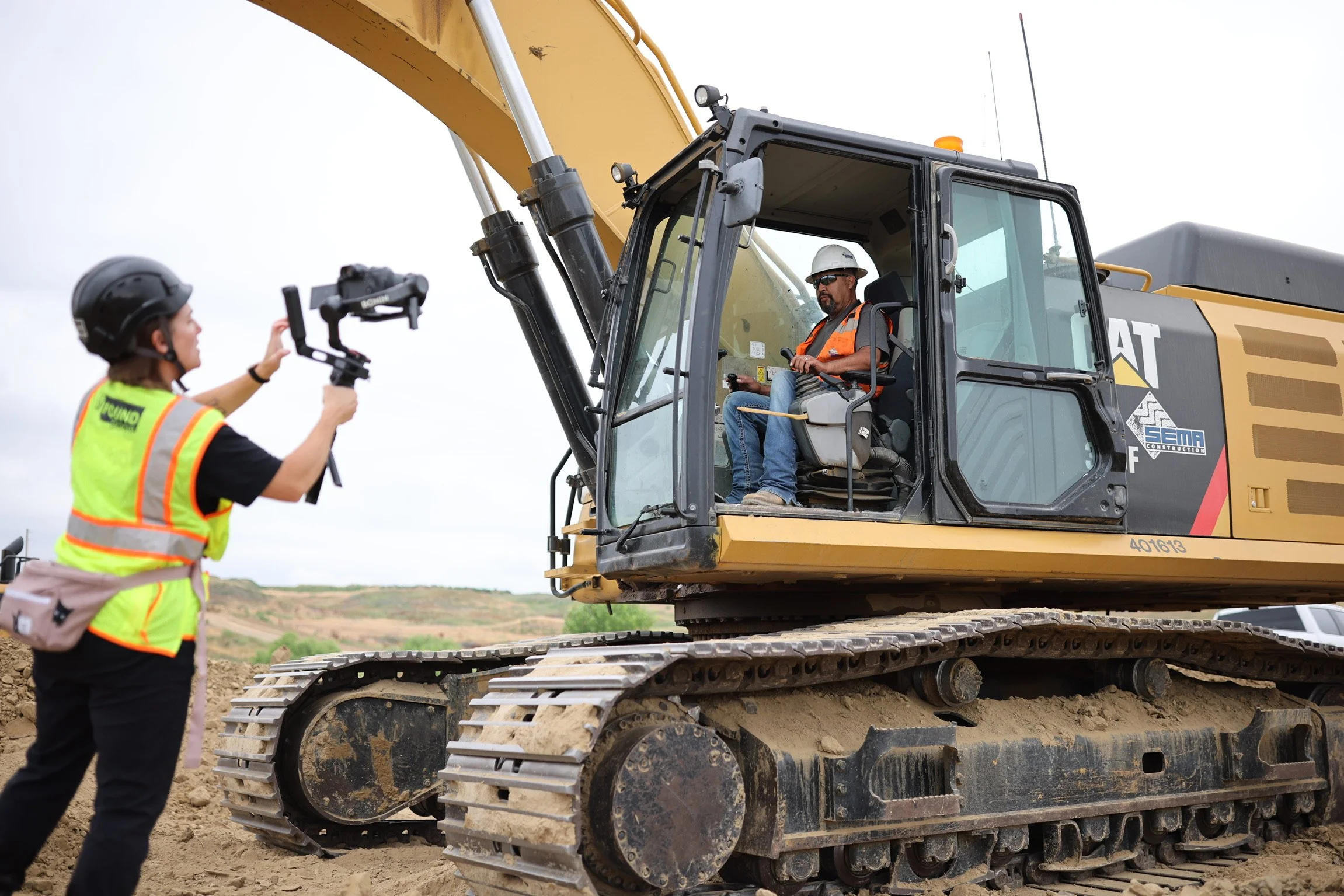 A woman in a safety vest and helmet filming a construction worker sitting inside an excavator with a camera stabilizer on a construction site during the daytime.