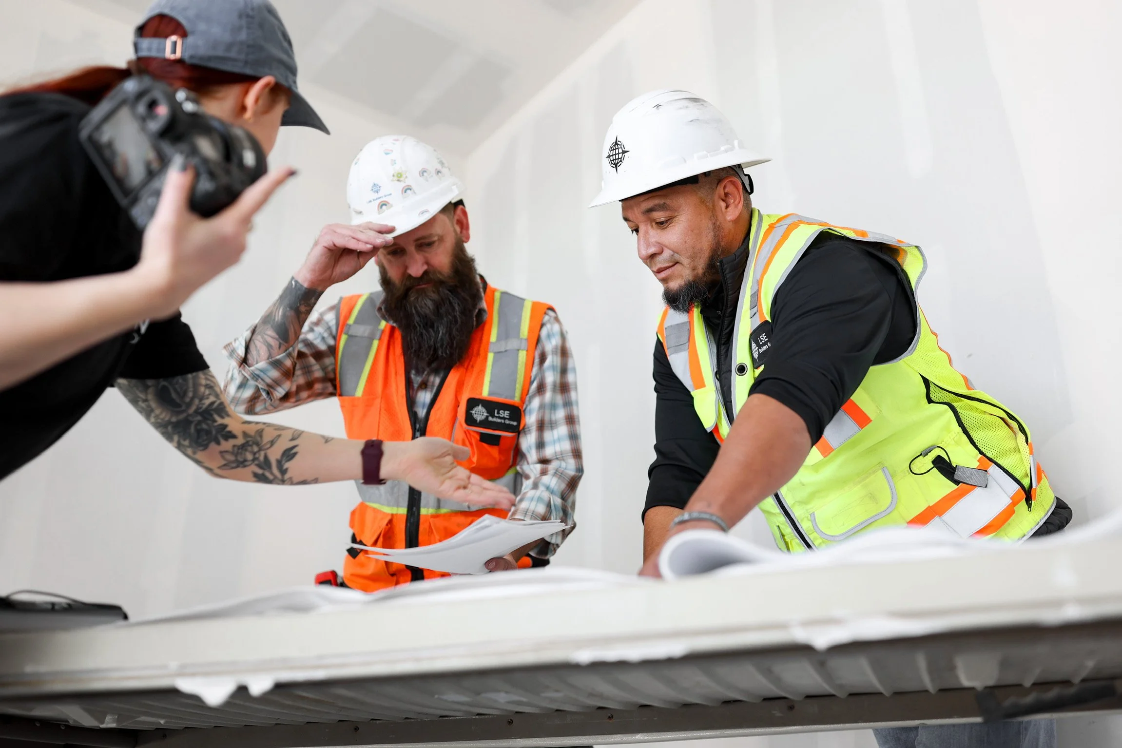 Construction workers reviewing plans with a woman holding a camera in an indoor setting.