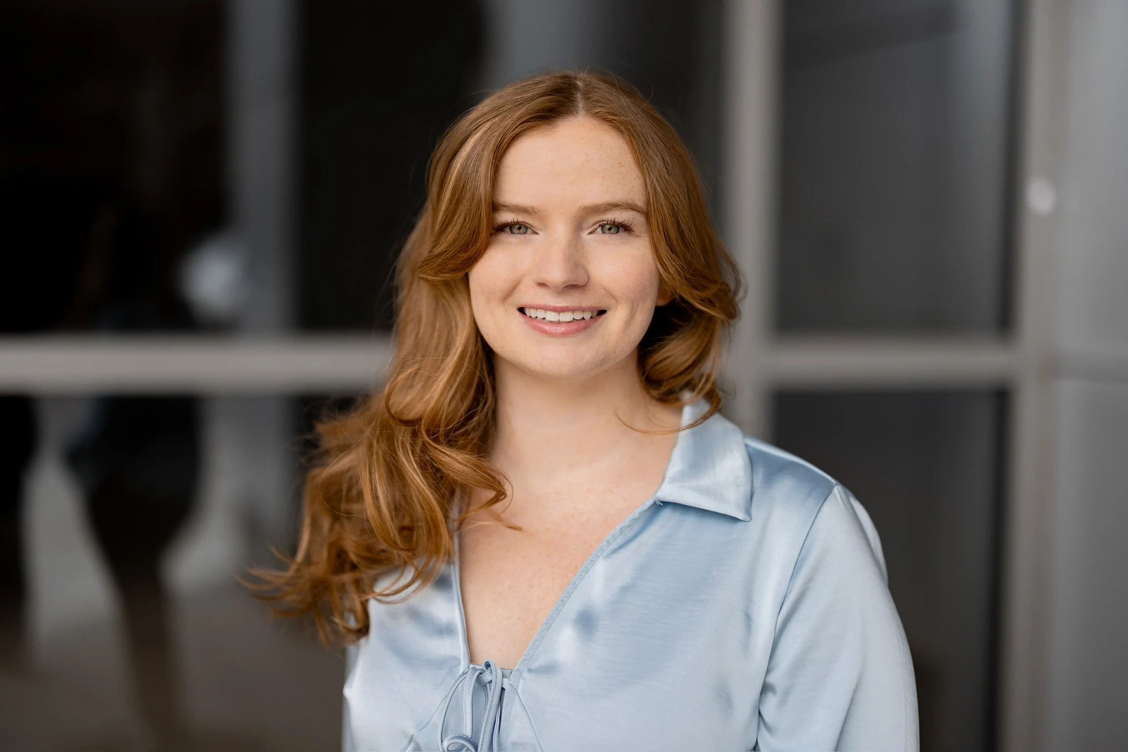A young woman with red hair and a light blue satin shirt smiling at the camera. She is in a modern indoor setting with blurred dark and light background elements.