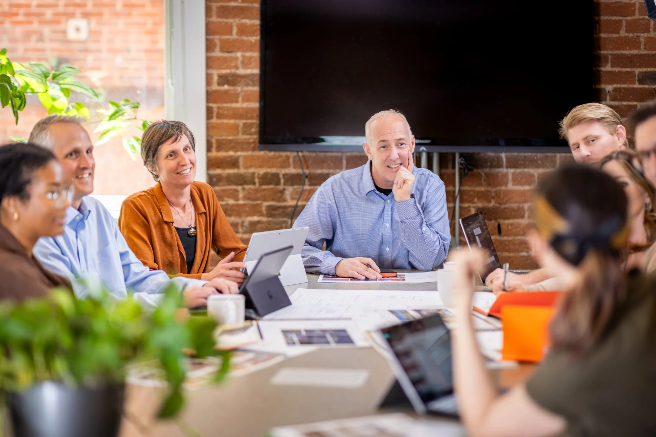 Group of professionals participating in a meeting in a modern conference room with exposed brick walls, laptops, tablets, notebooks, and coffee cups on the table, with a large TV screen on the wall.