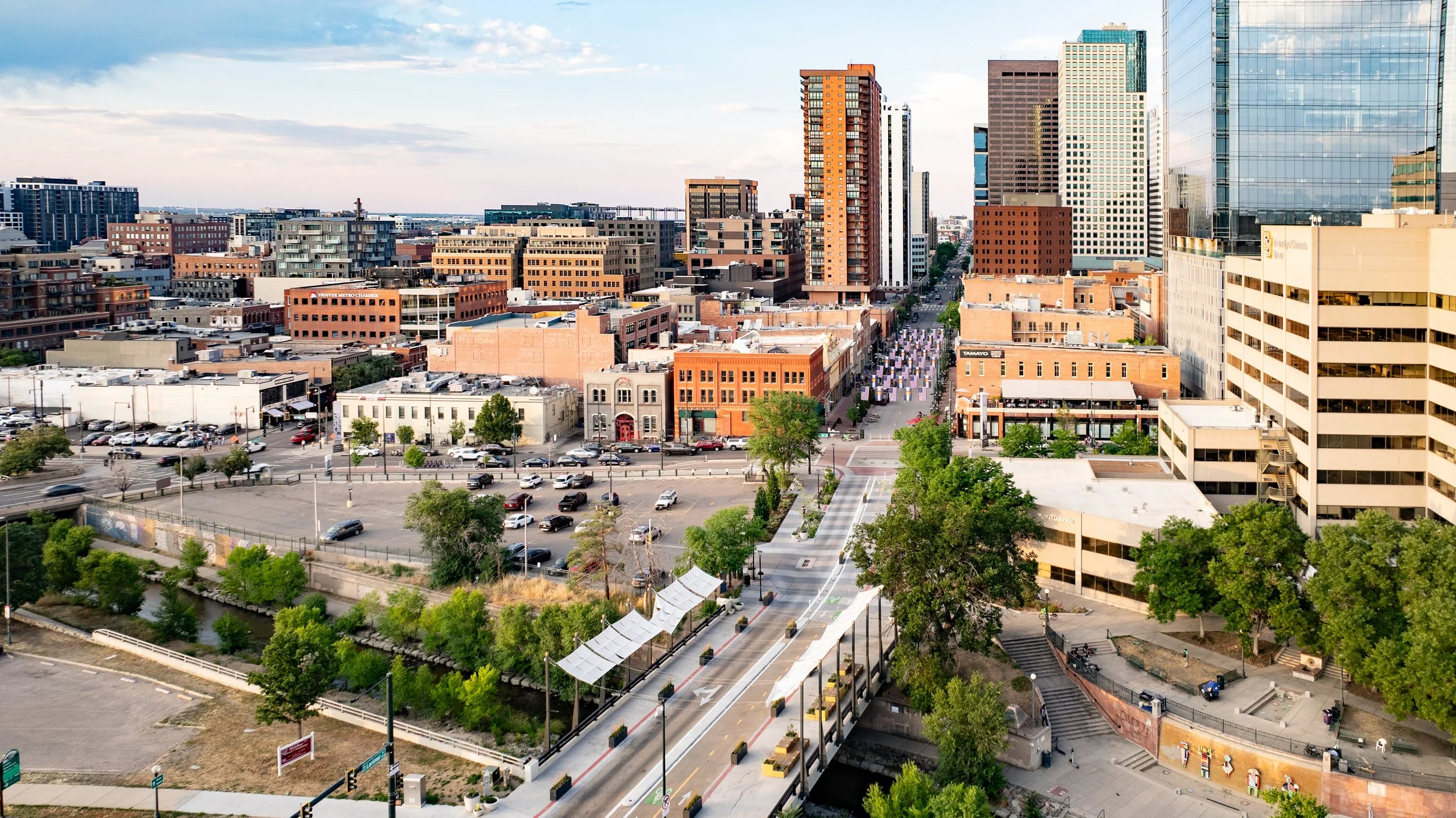 A cityscape with tall modern glass buildings and shorter brick buildings, a busy street with cars, trees, and people walking, a park with green trees and a creek in the foreground.