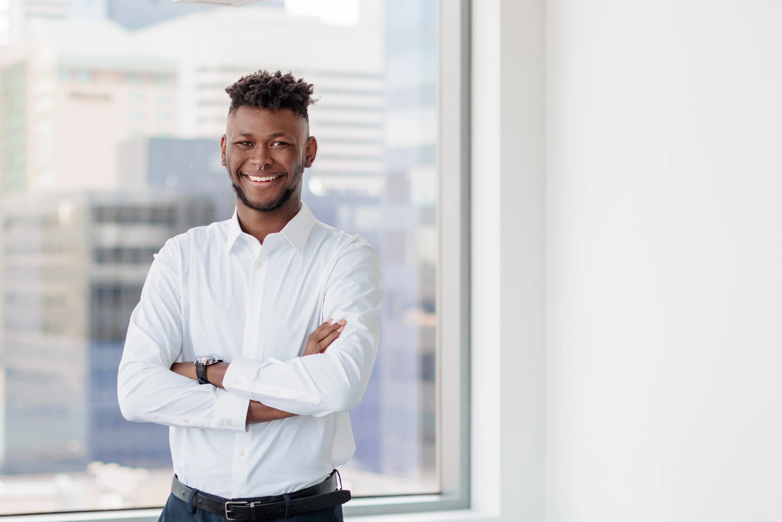 A smiling man with a beard and short curly hair standing with arms crossed in front of a large window in a modern office, wearing a white dress shirt and a watch.