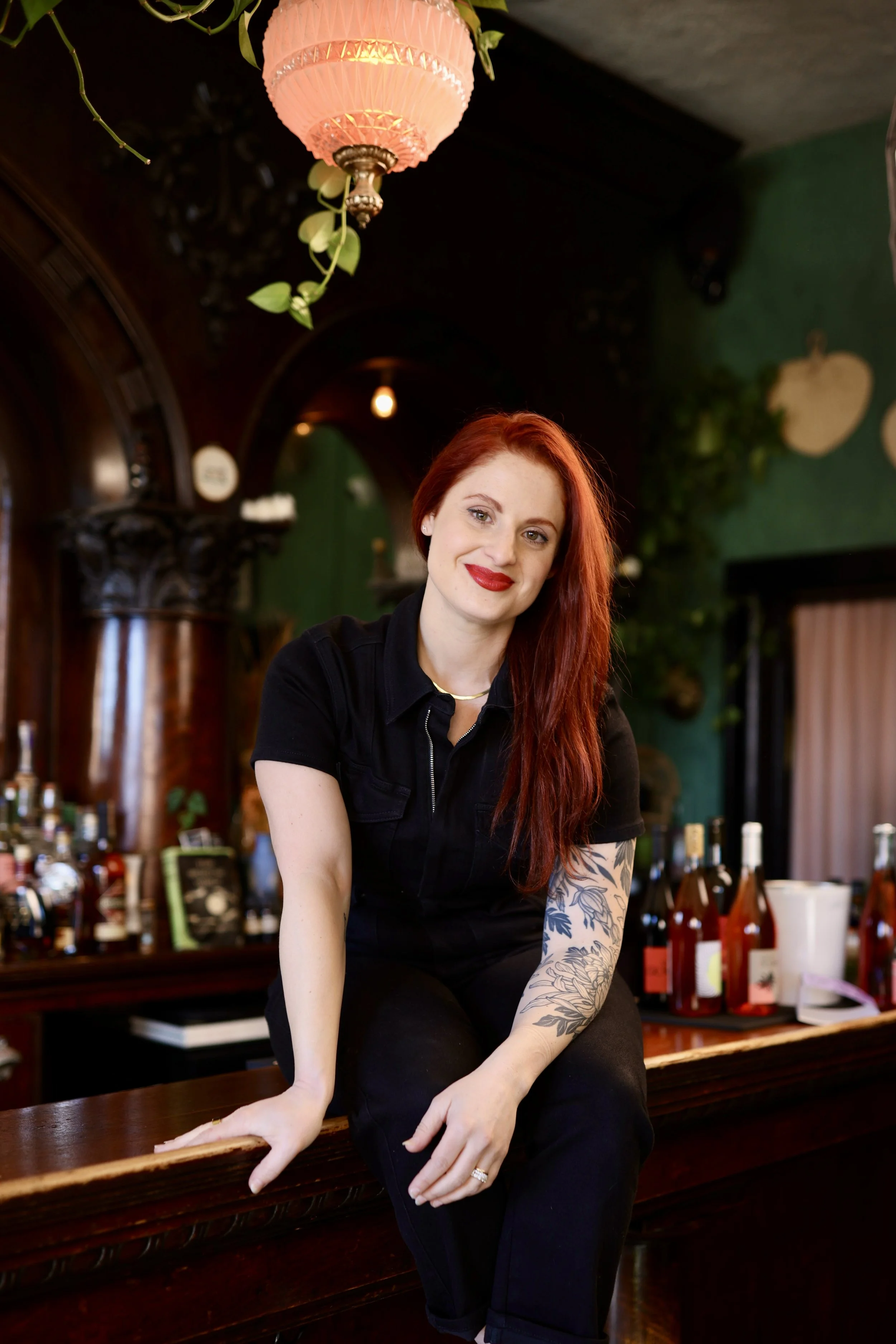 A woman with red hair and a tattoo on her arm, wearing a black shirt, sitting at a bar with bottles of alcohol behind her in a cozy, dimly lit setting with green walls and hanging lights.
