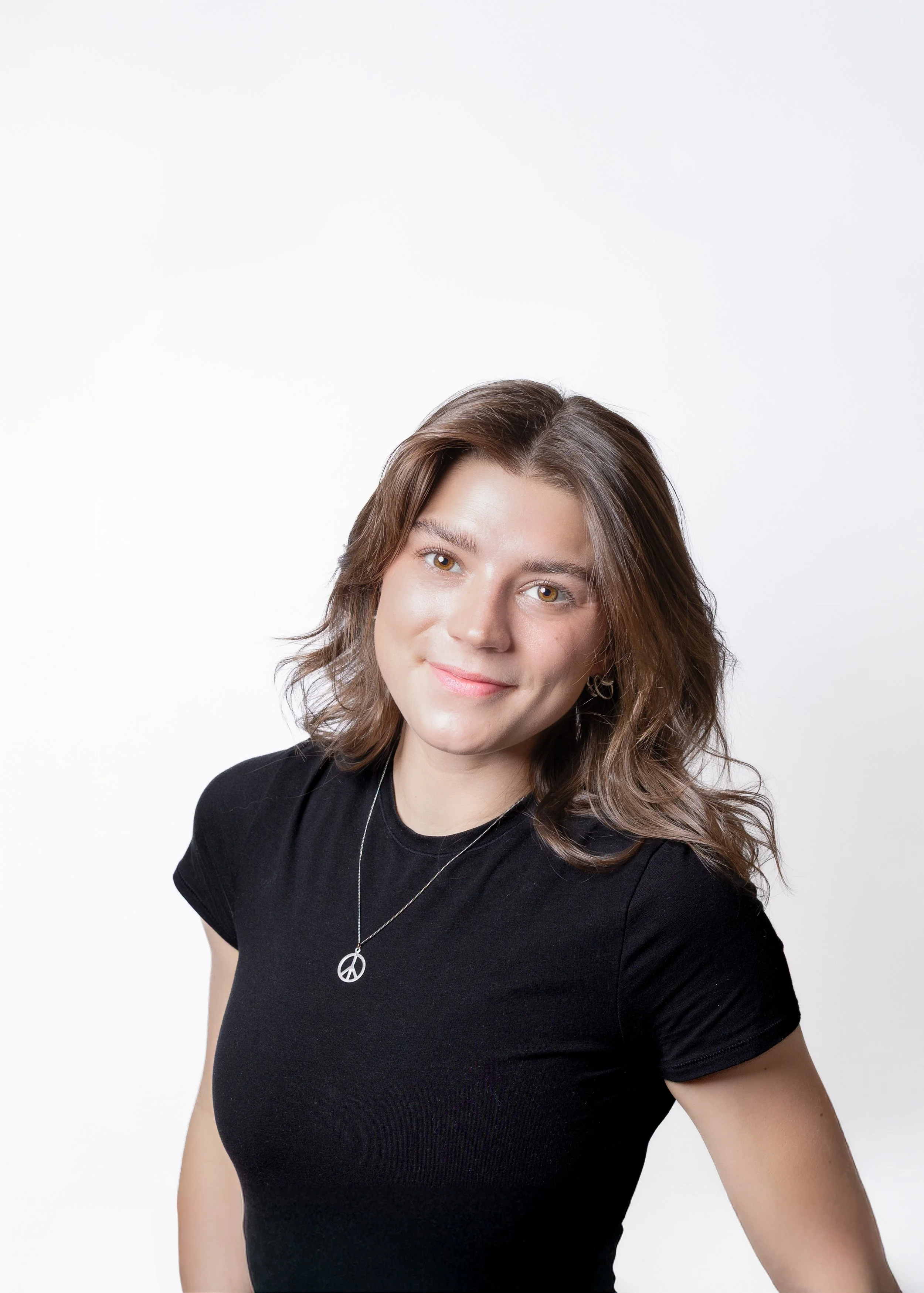 A woman with brown, wavy hair, wearing a black shirt and a peace sign necklace, smiling against a white background.