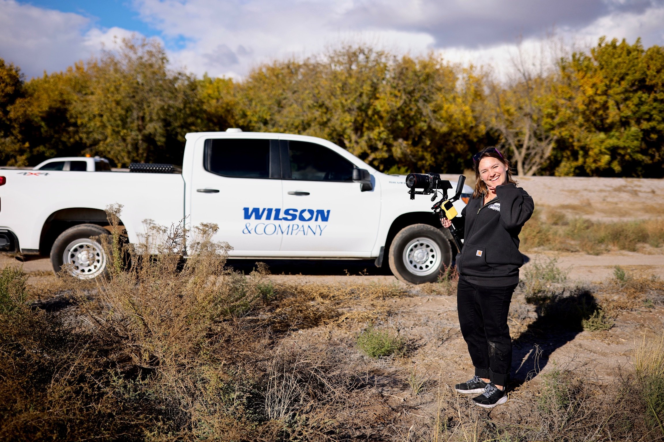 Woman smiling in outdoor desert landscape holding a professional camera gimbal with a camera attached, standing near a white Wilson & Company pickup truck, with an autumn-colored tree line in the background.