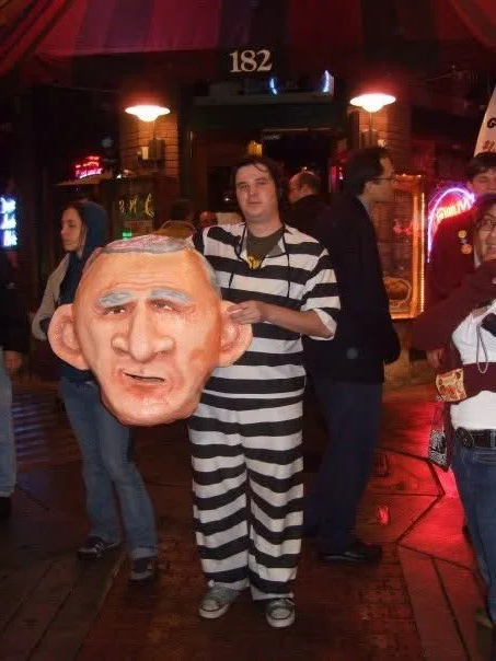 A young Thomas Wingate is wearing a prisoner costume and holding a giant costume head of George W Bush