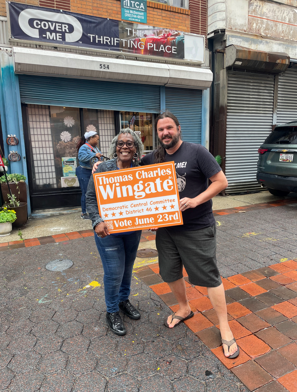 Two people standing on a sidewalk holding a sign promoting voting for Thomas Charles Wingate in District 46, with a storefront and people in the background.