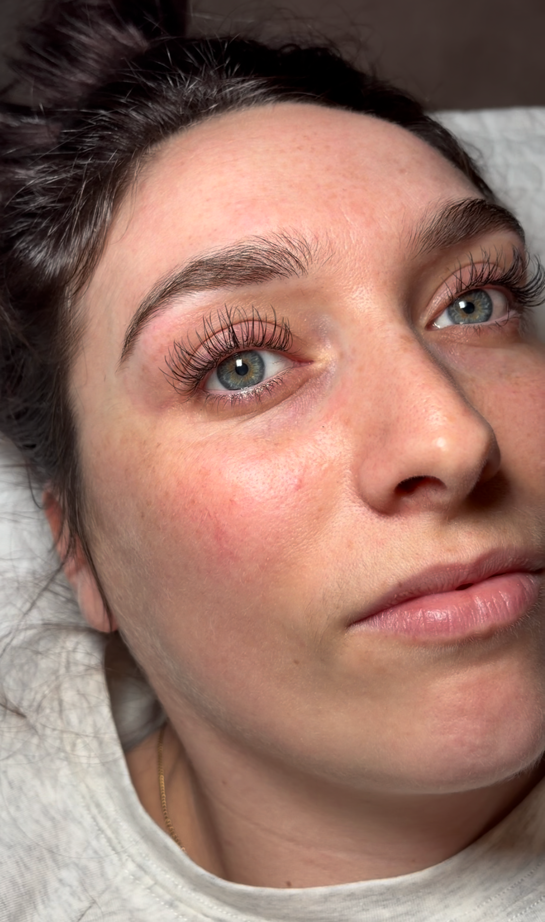 Close-up of a woman with blue eyes, natural makeup, and long curled eyelashes lying down on a pillow.