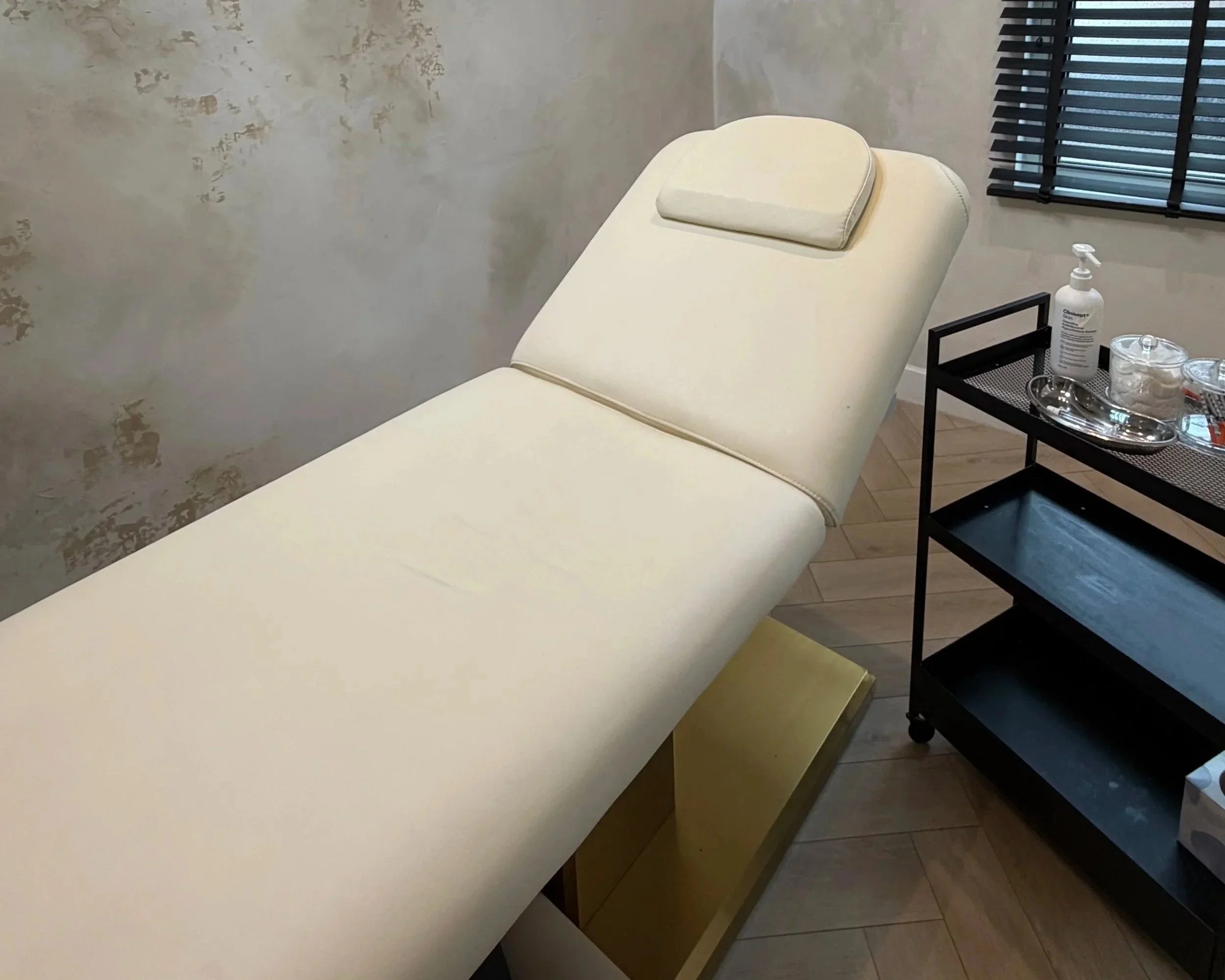 Treatment bed with head cushion, situated in a room with textured wall, wooden floor, and black window blinds.