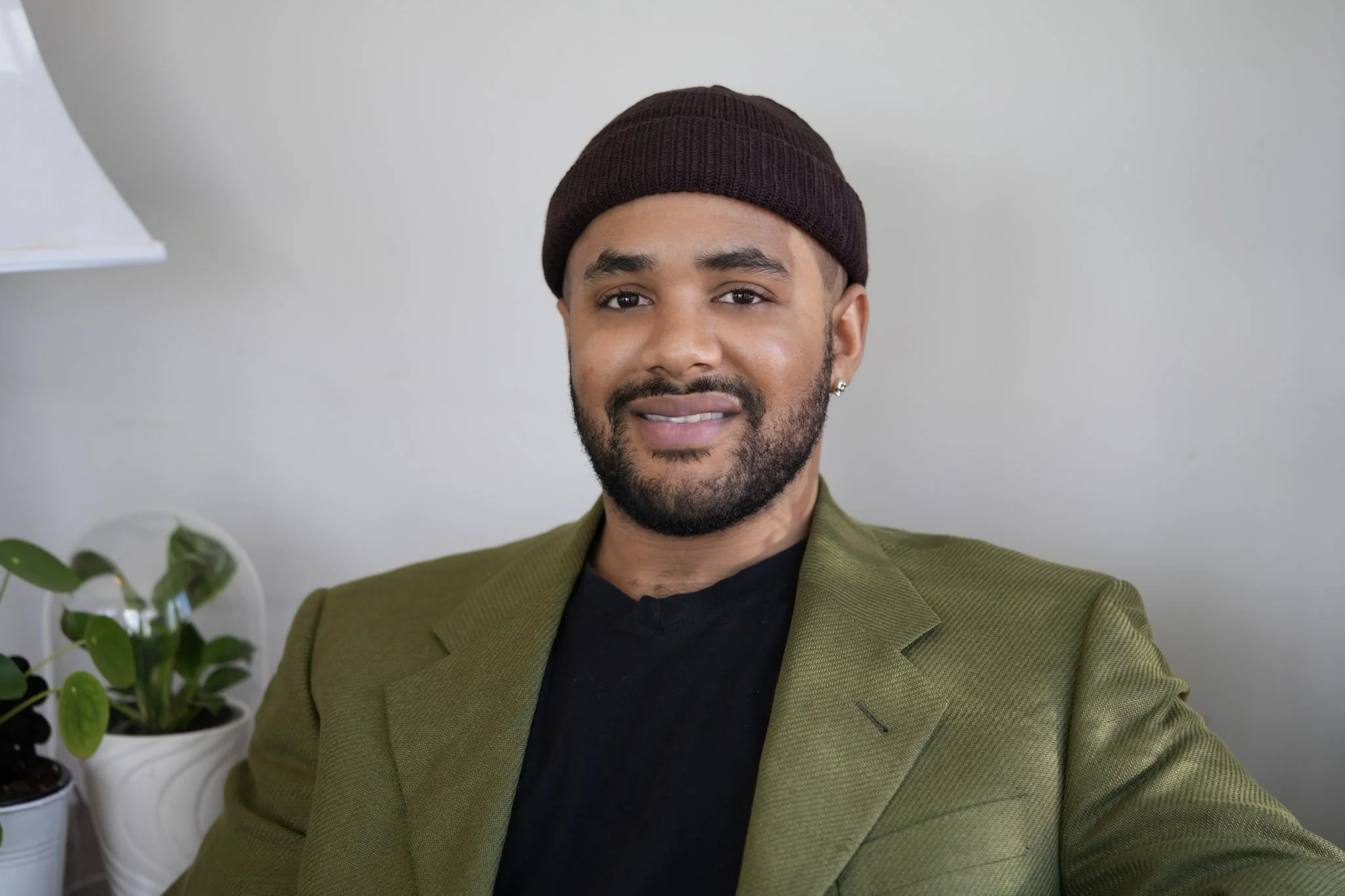 A man with a beard, wearing a brown beanie, black shirt, and olive green blazer, sitting indoors with a neutral background and a potted plant visible in the background.