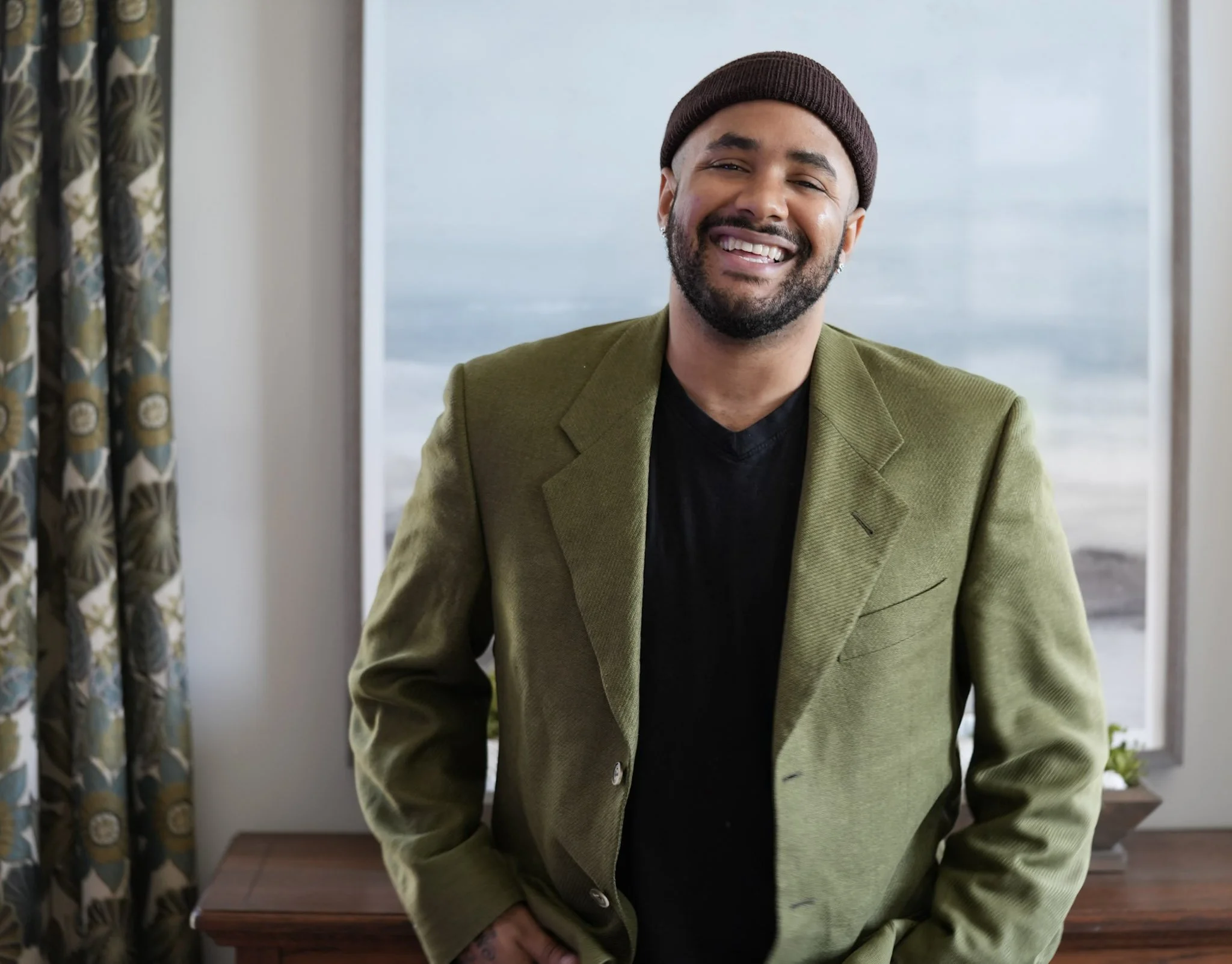 A smiling man with a beard and mustache wearing a brown beanie, a green blazer, and a black shirt, standing indoors near a window, with patterned curtains on the left and a piece of wooden furniture below the window.