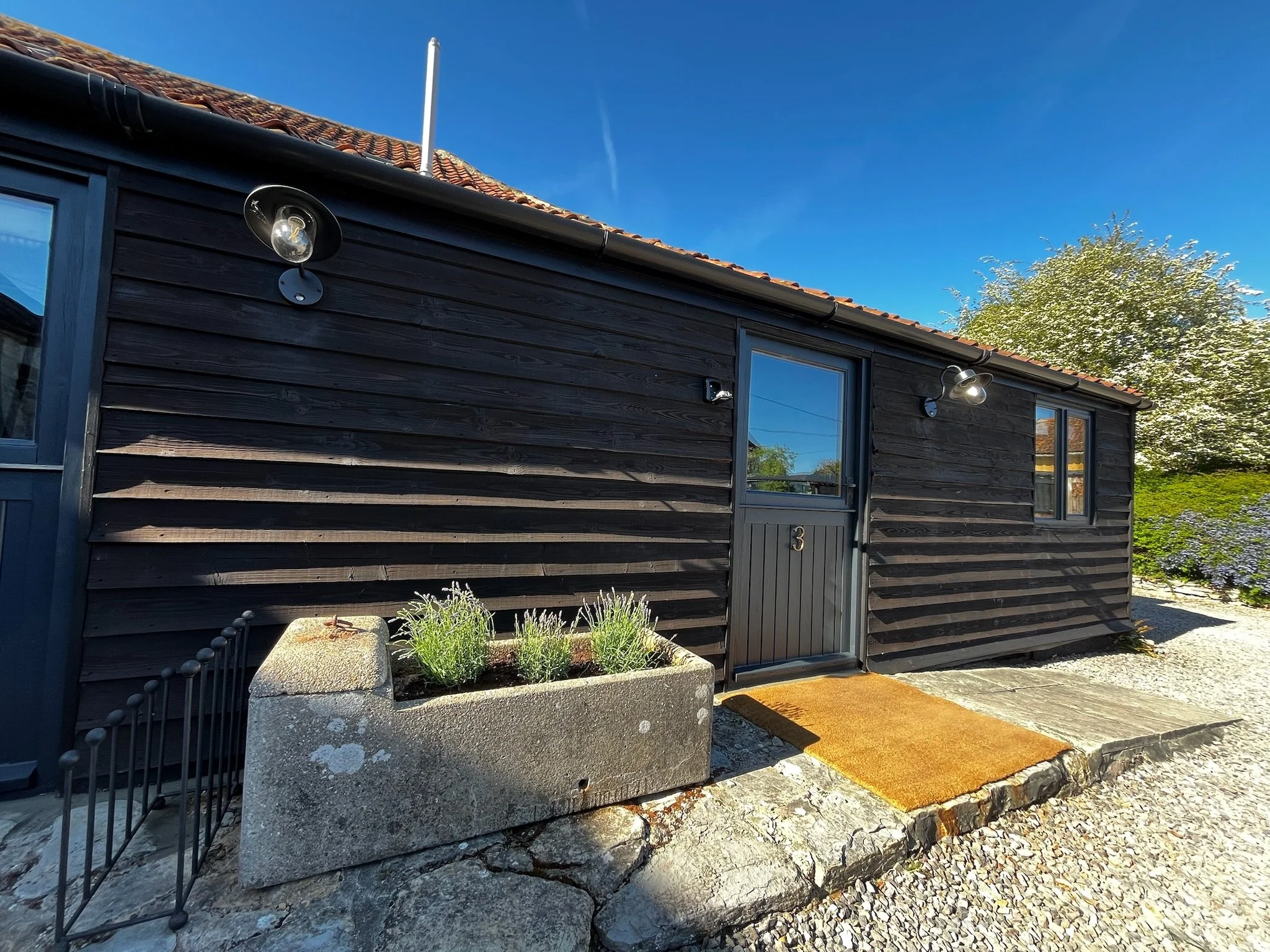 A modern house exterior with dark wooden siding, large windows, and a front door. There are two wall-mounted outdoor lights and a small garden bed with plants. The house has a tiled roof and a clear blue sky in the background.